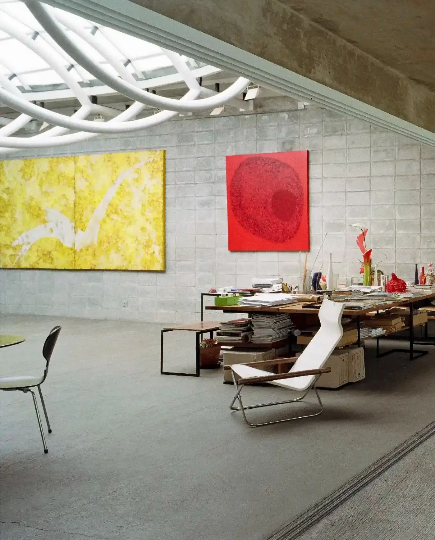 Sala de trabalho com paredes de blocos de concreto, contendo duas obras de arte abstratas, uma amarela e uma vermelha, além de uma mesa com muitos papéis e objetos diversos, uma cadeira de descanso branca ao lado.