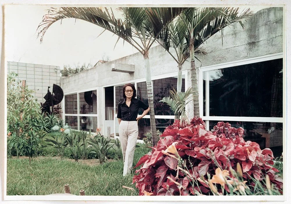 Tomie Ohtake, mulher de cabelos castanhos, usando blusa preta e calças claras, de pé em jardim com plantas, árvores e plantas tropicais ao lado de uma casa moderna com janelas grandes.