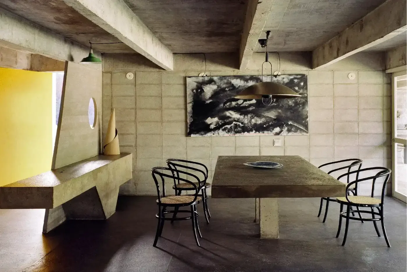 Sala de jantar moderna com parede de blocos de concreto, quadro abstrato preto e branco, mesa de madeira com cinco cadeiras de vime, janela grande e iluminação suave com dois pendentes de metal.