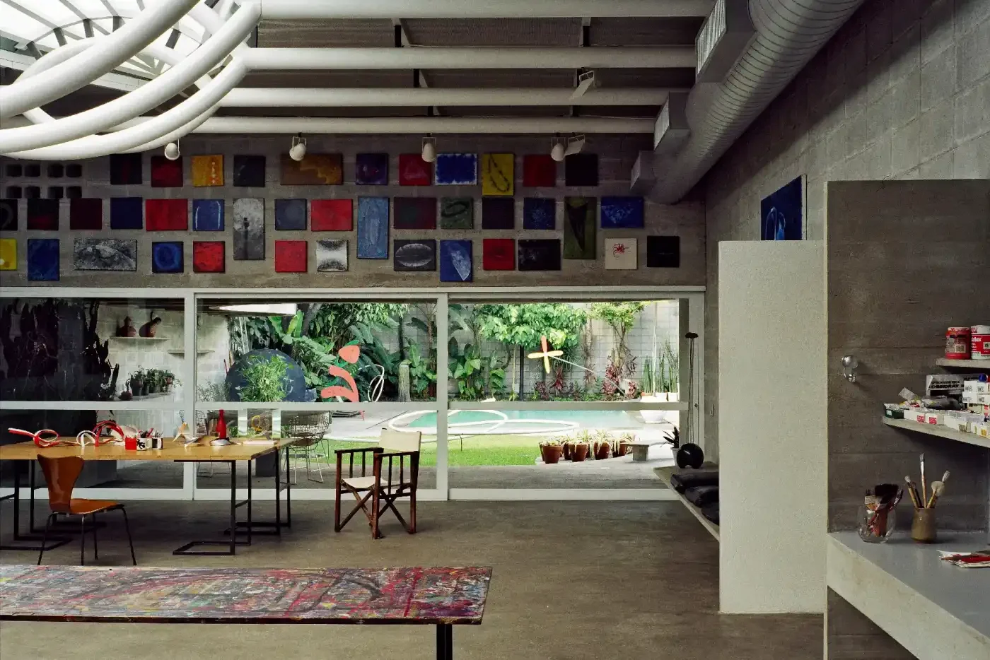 Interior de uma sala com parede de vidro, mesas, cadeiras, obras de arte nas paredes e no teto, jardim com plantas e piscina ao ar livre.