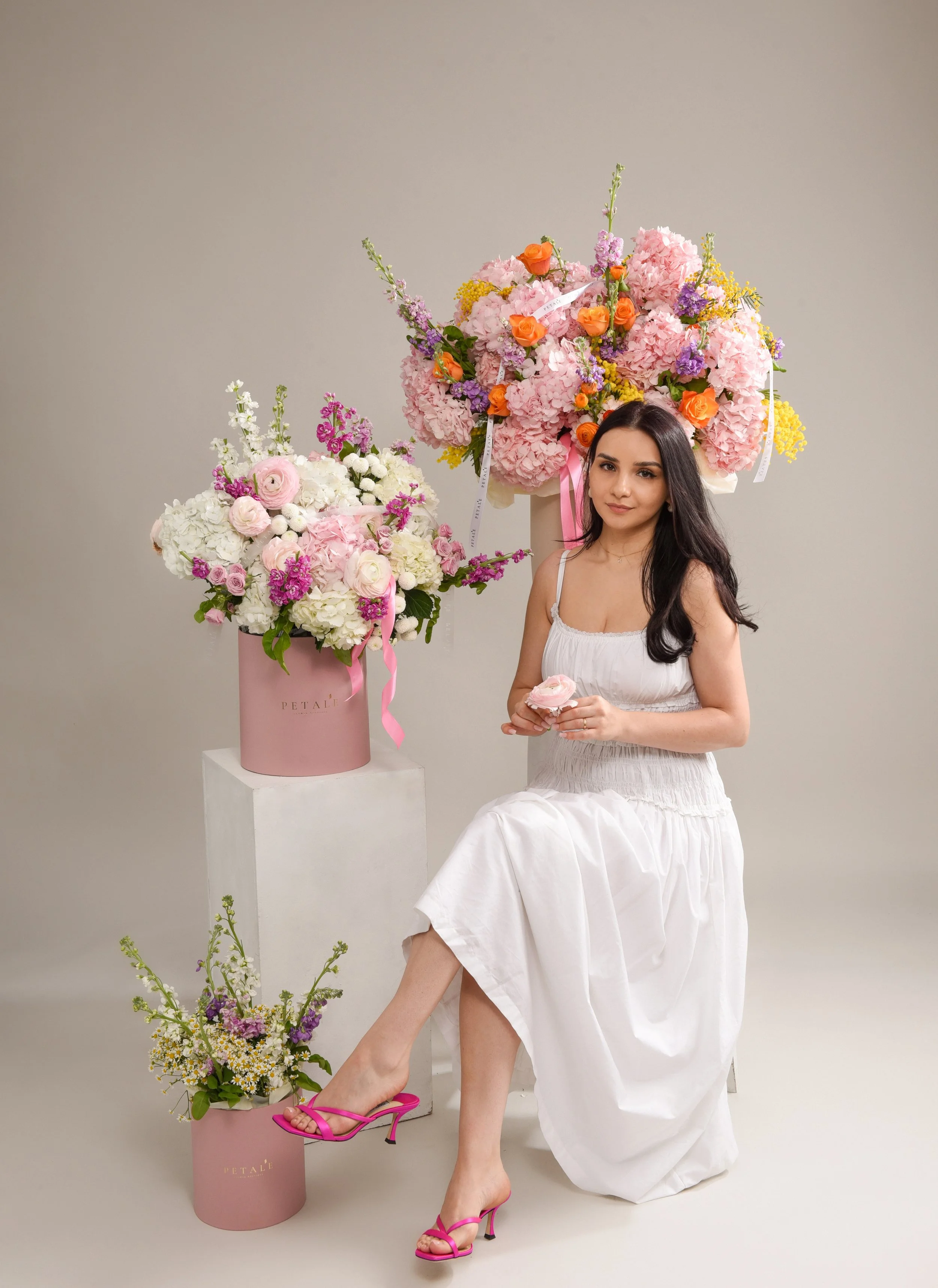 A young woman in a white dress sitting in front of pink and white floral arrangements, holding a pink flower.  Florist photoshooting ideas. Aesthetic flower shop Manchester