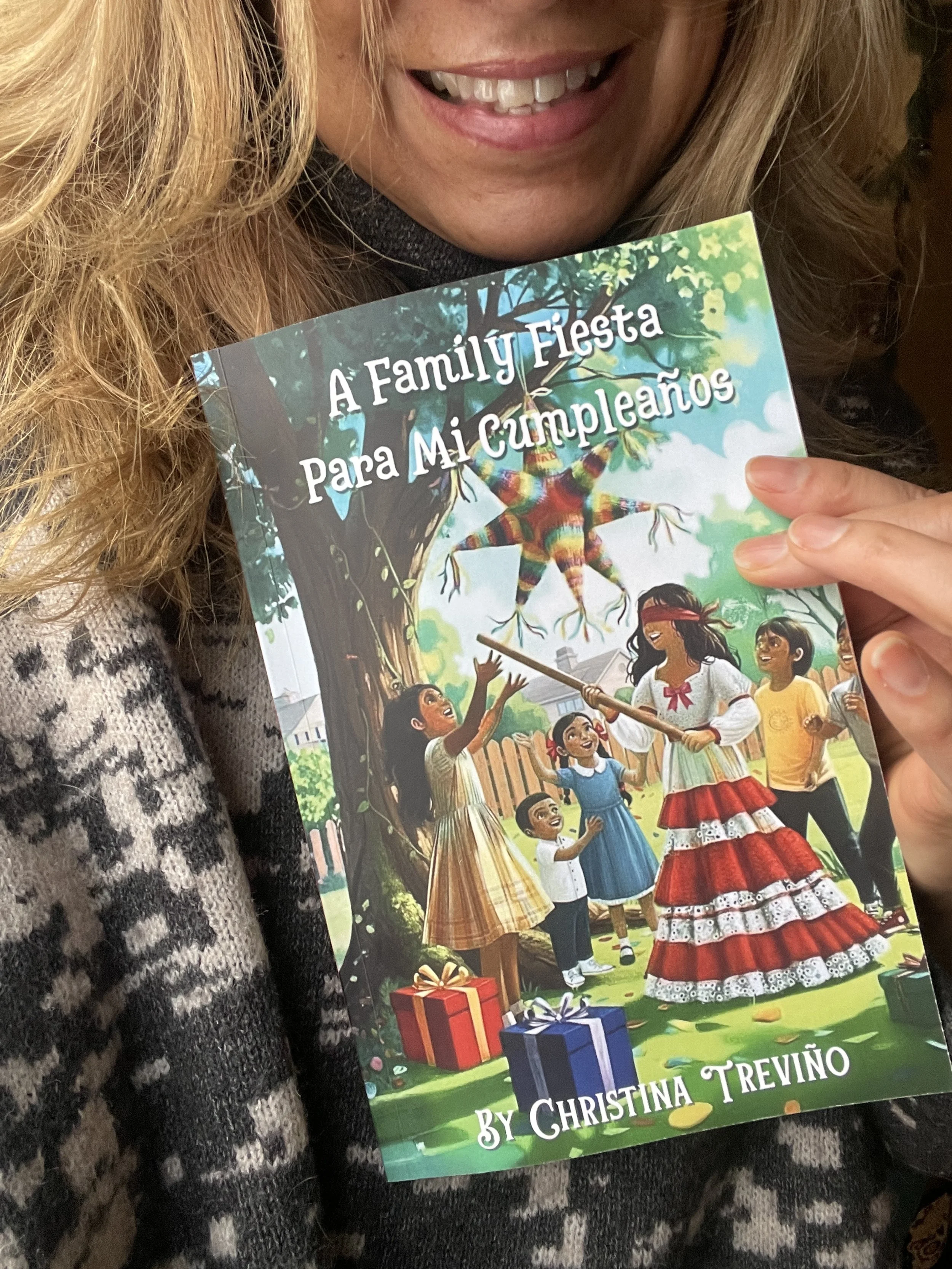 A woman with blond hair smiling and holding a colorful children's book titled "A Family Fiesta Para Mi Cumpleaños" by Christina Treviño. The cover illustration depicts children celebrating outdoors with presents and a girl dressed as a piñata.