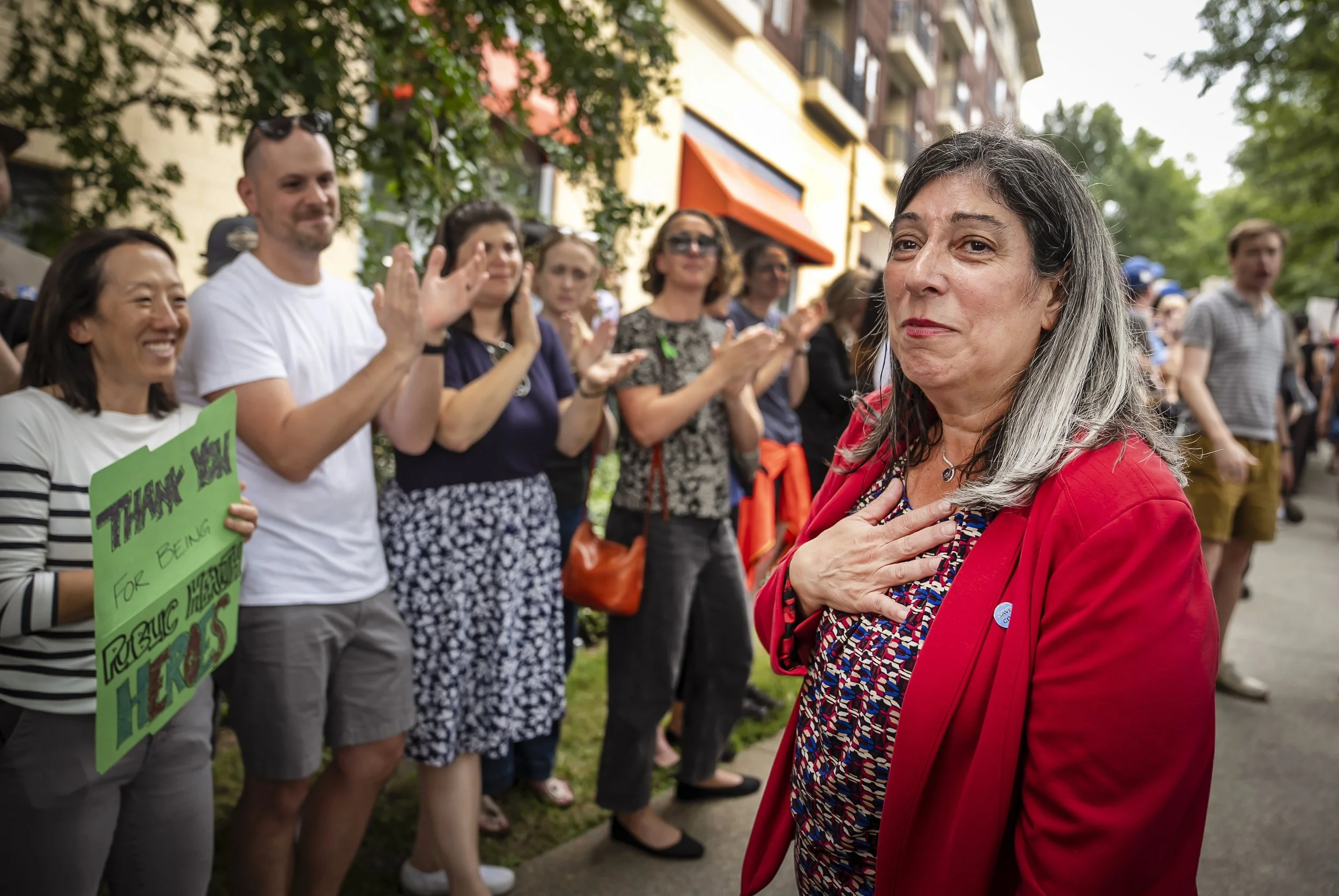 A woman with gray hair and a red jacket standing with her hand over her heart, while a group of people behind her are clapping and holding a sign that says "Thank you for being true to yourself." in a line outdoors.