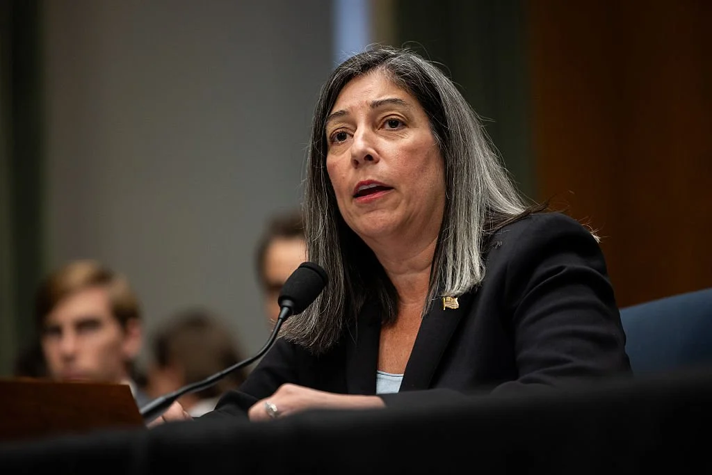 A woman with long gray hair speaking into a microphone at a formal event or hearing.