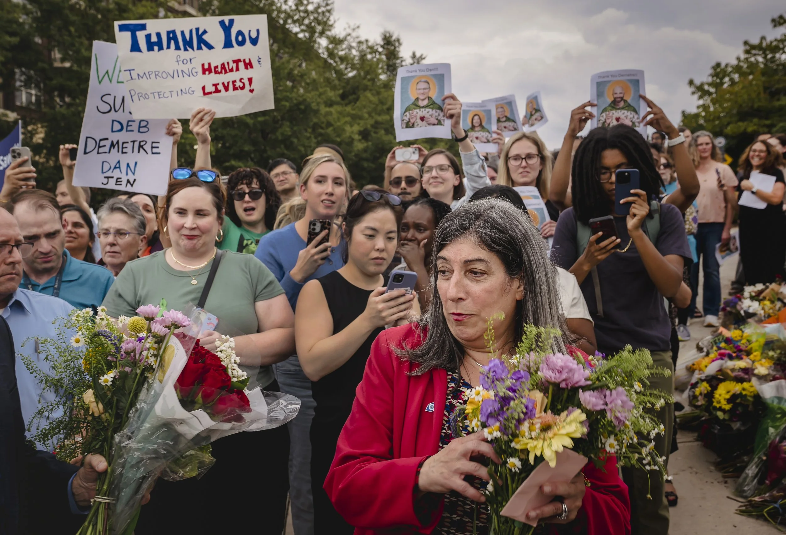 A woman in a red jacket holding a bouquet of flowers is at a gathering of people, some of whom are holding signs and taking photos or videos. The signs express gratitude for healthcare workers. The crowd is diverse and appears to be attending a memorial or tribute event, with flowers laid out on the ground.