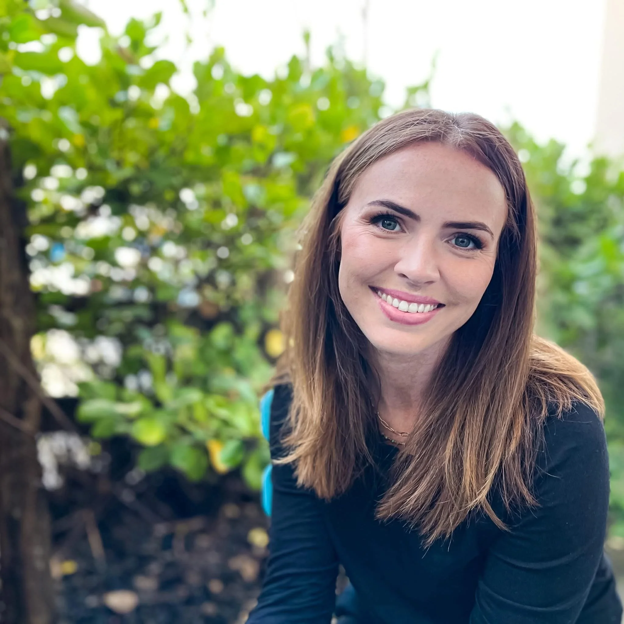 A woman smiling outdoors with a background of green leaves and blurred greenery.