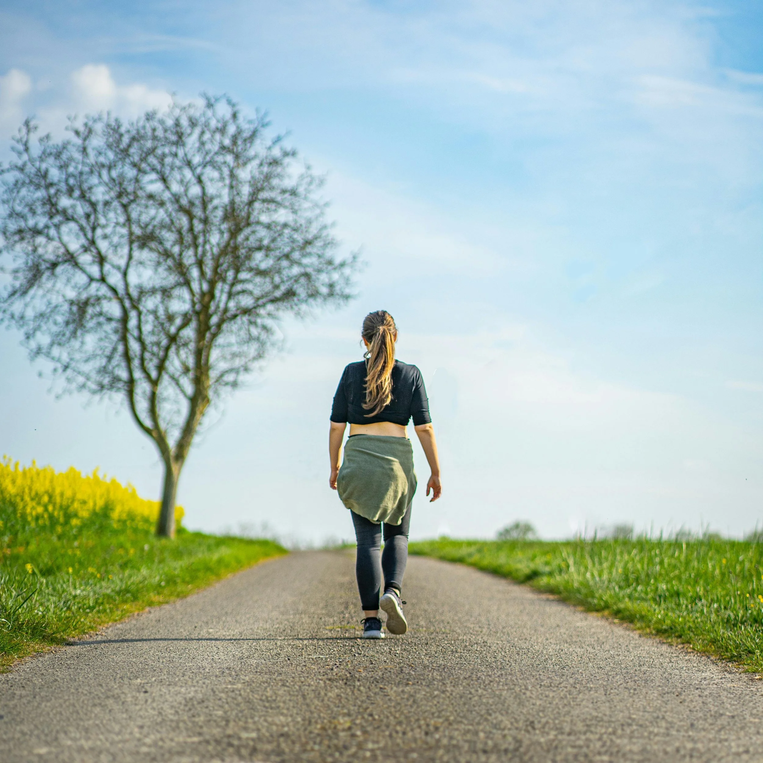 A woman walking on a paved country road during the day, with green grass and yellow wildflowers on the sides and a leafless tree on the left, under a partly cloudy sky.