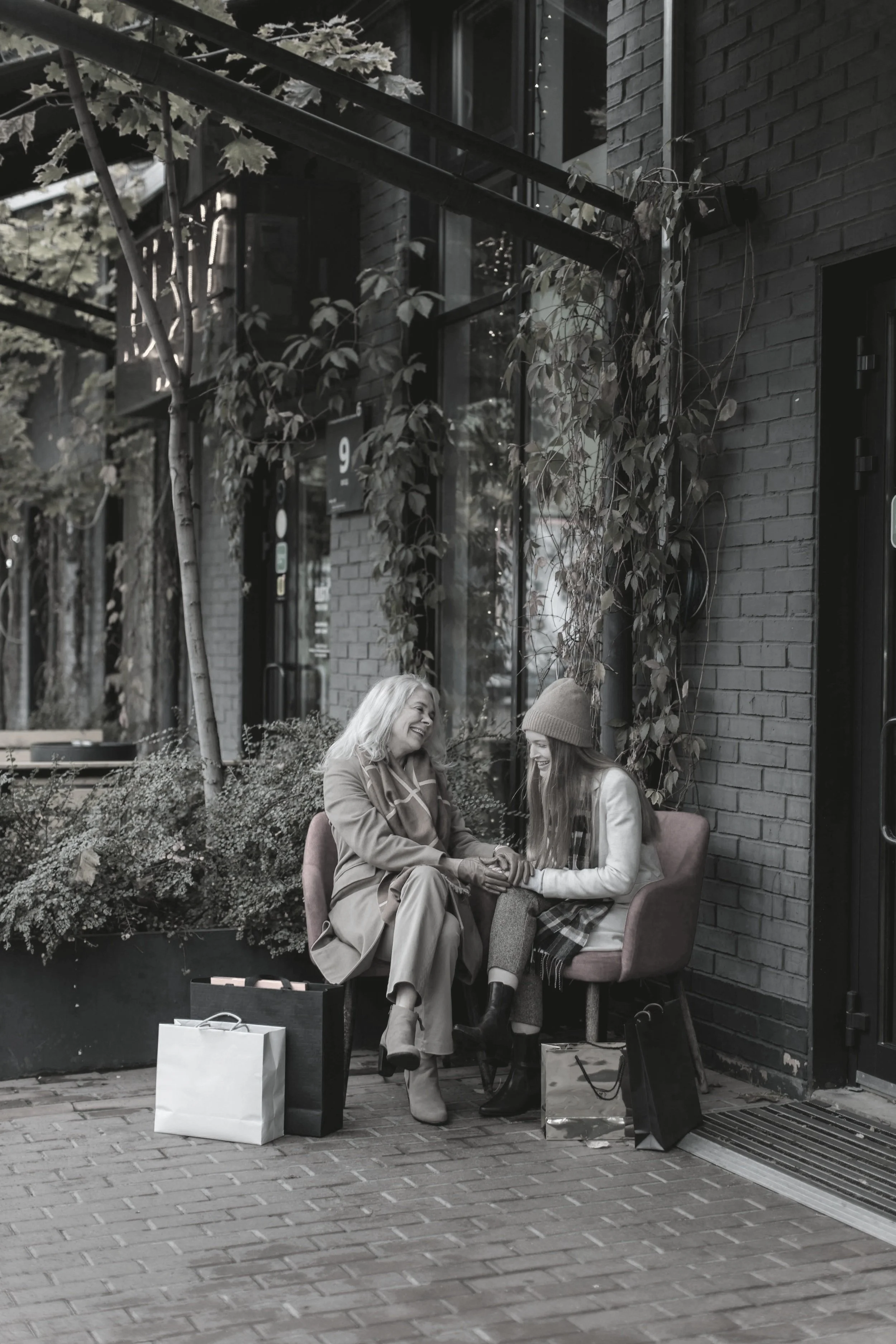Two women sitting outside on a bench, holding hands and smiling, surrounded by shopping bags, with a brick building and plants in the background.