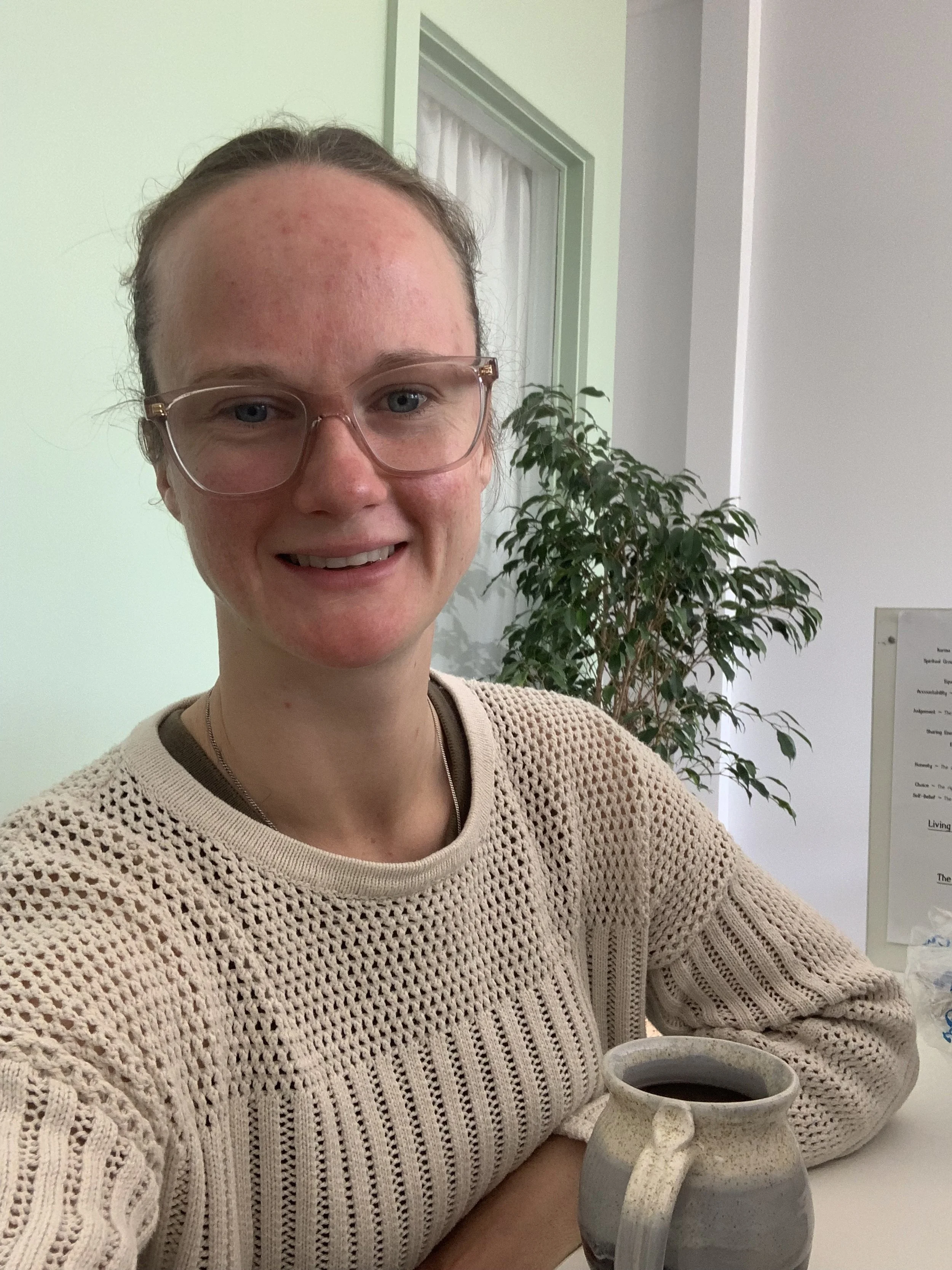 A woman wearing a cream jumper and glasses, holding a mug, with an indoor plant in the background.