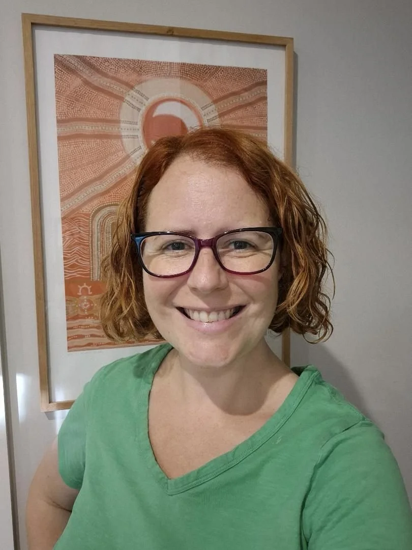 A woman wearing a green t-shirt and glasses with an orange indigenous print hanging on the wall behind