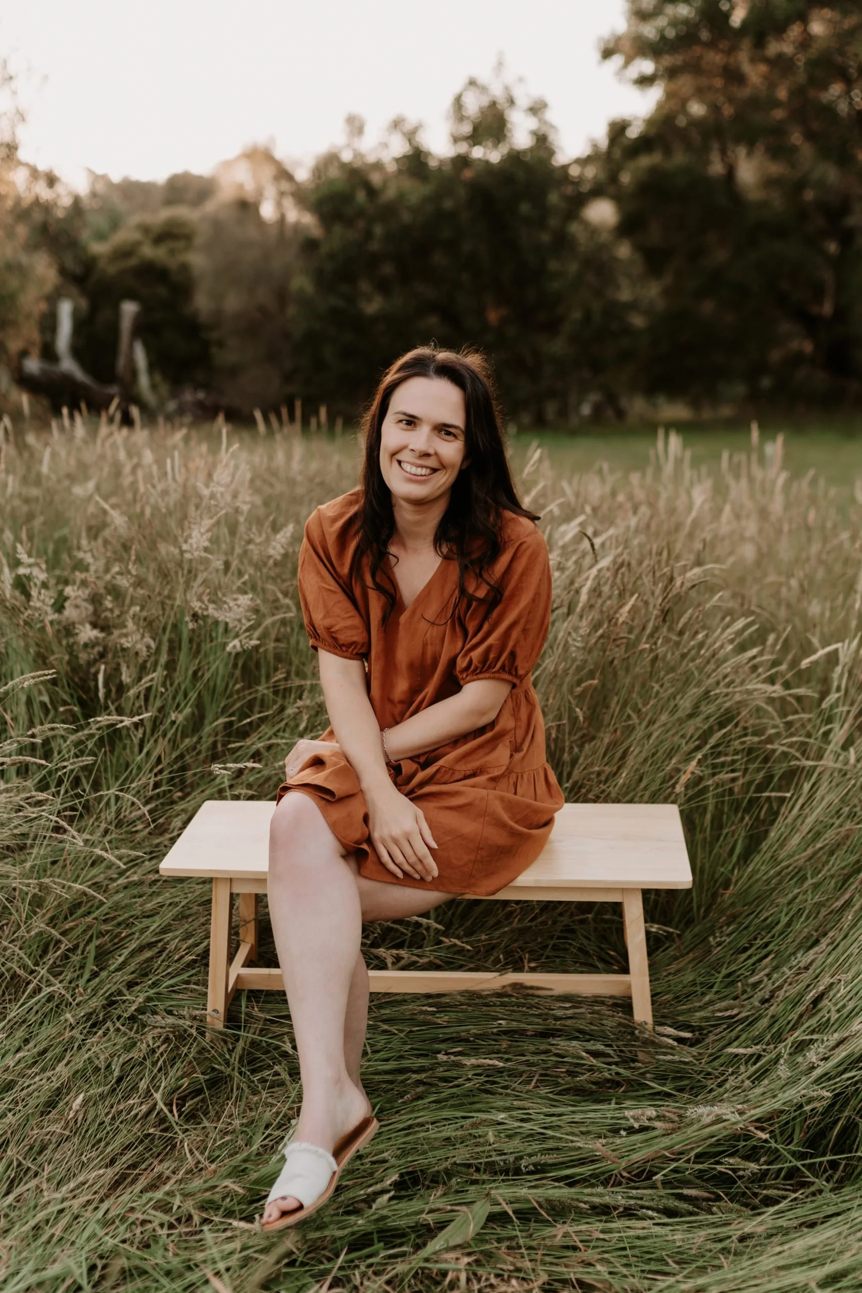 A woman with long dark hair sitting on a wooden bench with her legs crossed looking at the camera; in a field with long yellow and green grass