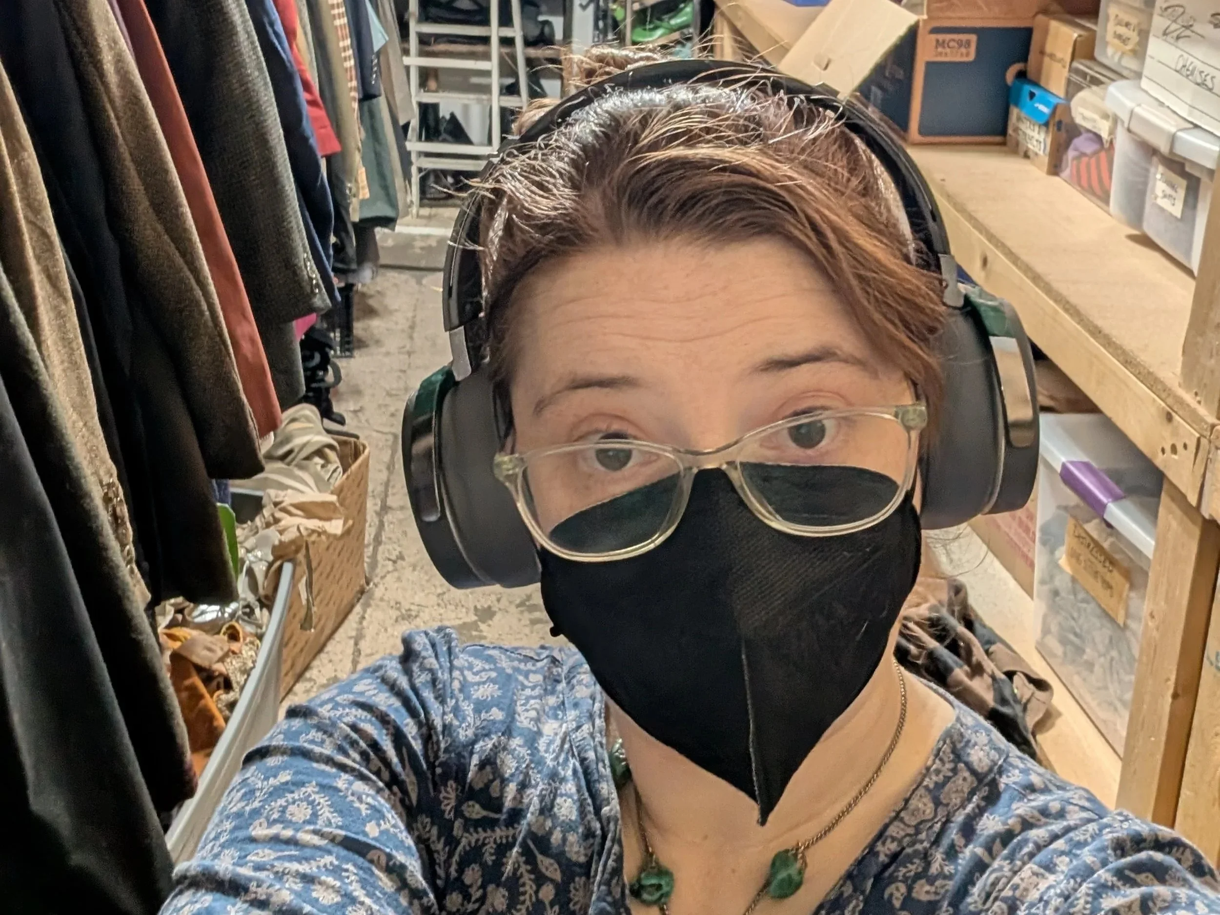 woman with red hair in a black kn95 mask, glasses, and headphones in a selfie standing in front of a messy costume storage