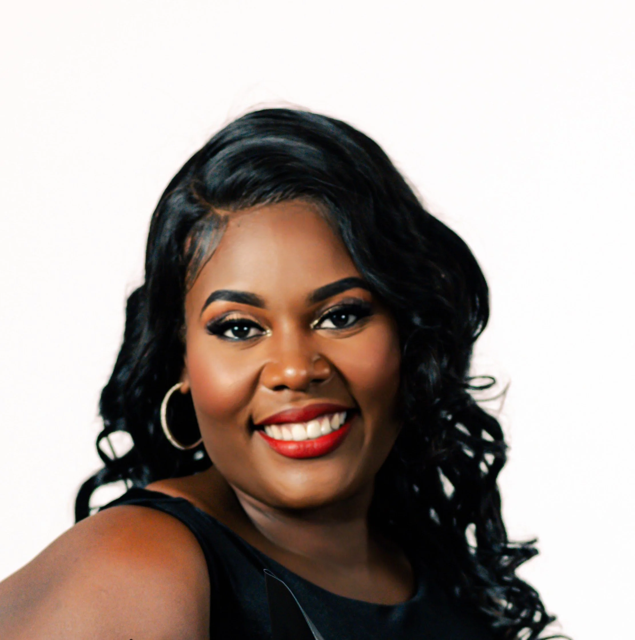 Close-up portrait of a woman with dark, wavy hair, red lipstick, and hoop earrings, smiling against a white background.