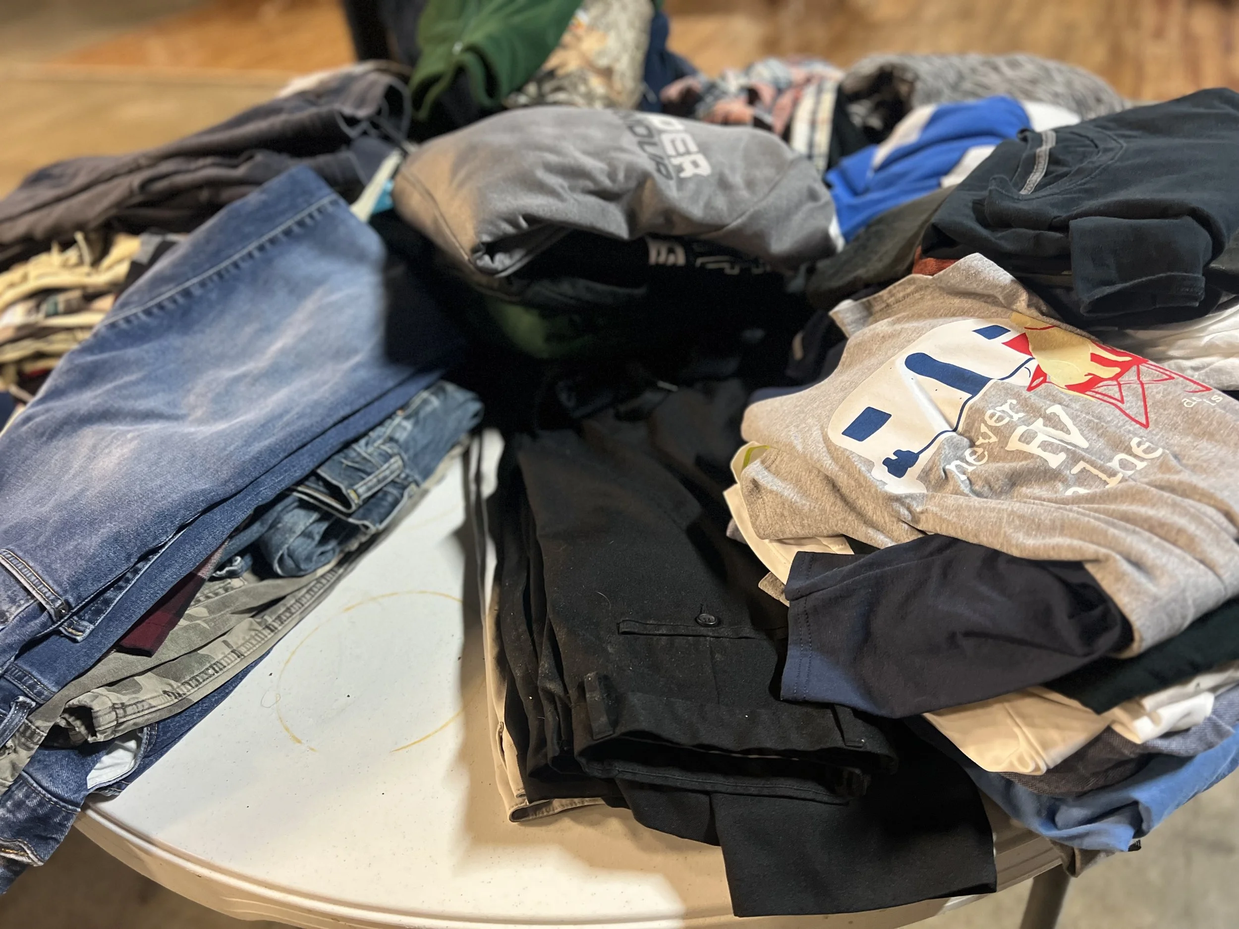 A large pile of folded clothes and t-shirts on a white table, including jeans, t-shirts, and other apparel.