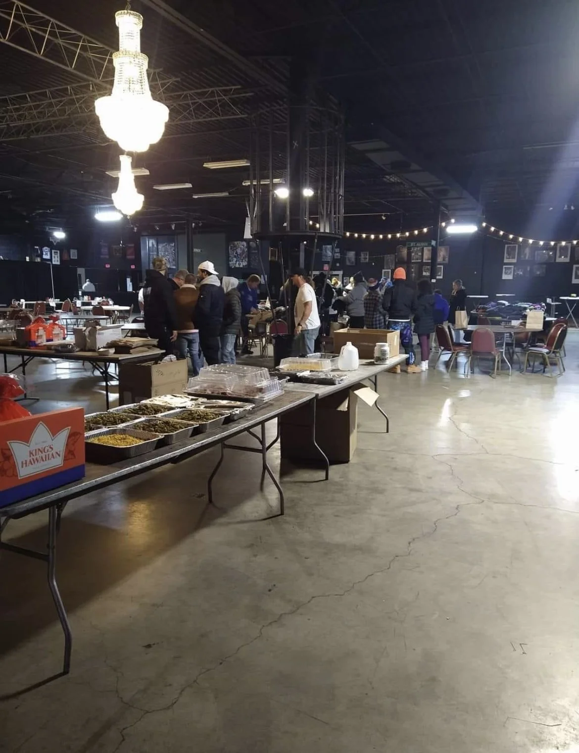 People gather around tables with food and drinks in a large indoor space with high ceilings and chandeliers.
