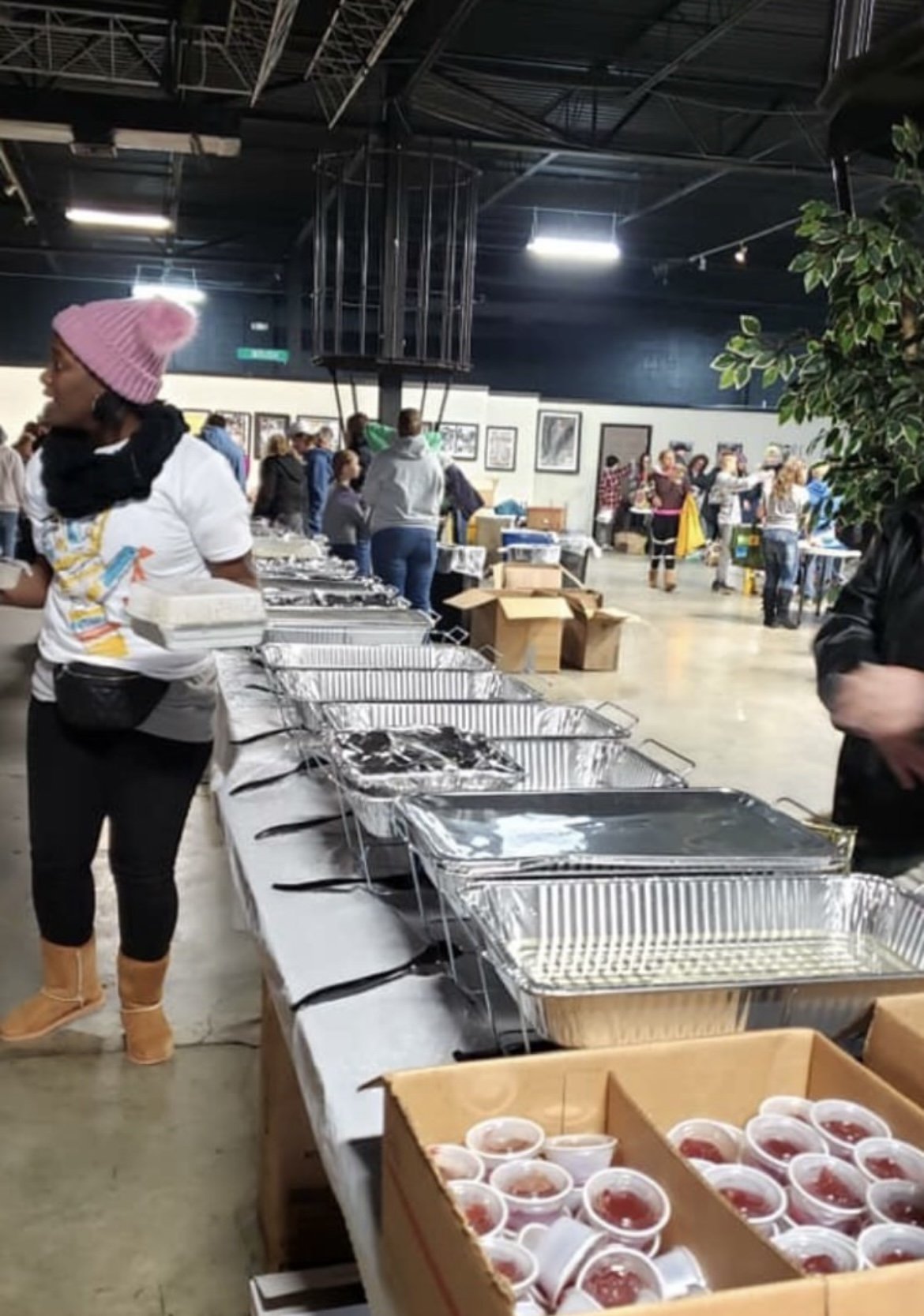 People serving and browsing food at an indoor food event or market with tables, boxes, and trays of food, including desserts, in a spacious, industrial-style venue.