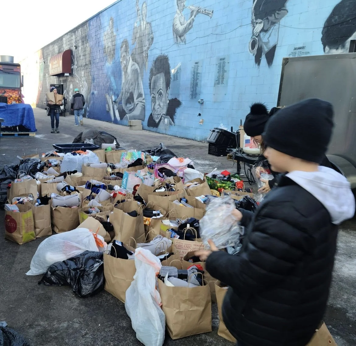 People organizing and distributing supplies in an outdoor area next to a mural painted on a building wall, with table and tires in the background.