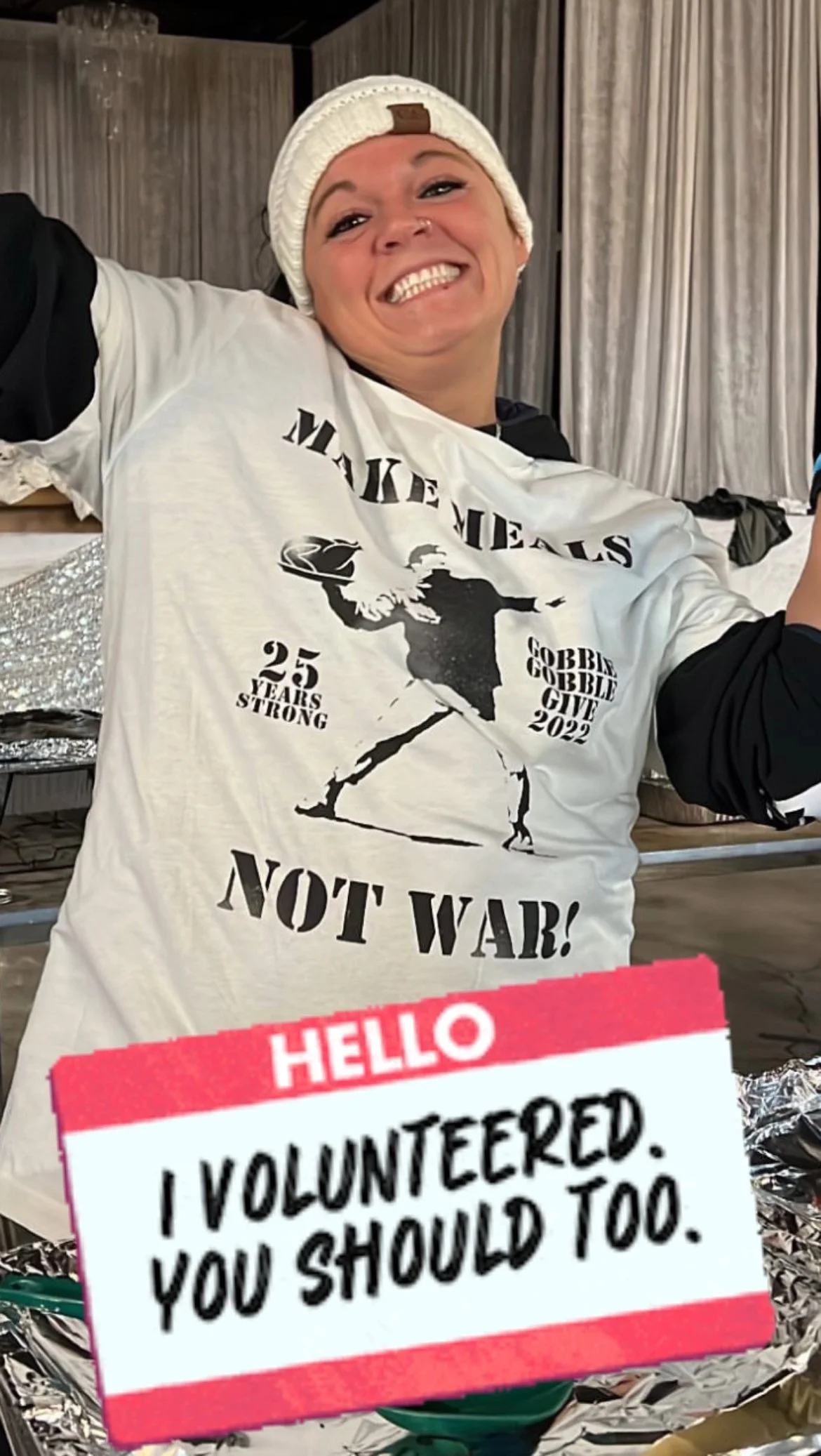 A woman smiling, wearing a white knit cap and a white T-shirt with black text and graphics, standing indoors. The T-shirt features a snowboarder silhouette, and phrases like "Make Meals," "25 Years Strong," "Gobbler Gobble Give 2022," and "Not War!". There is a pink and white sign reading "HELLO I VOLUNTEERED. YOU SHOULD TOO." in front of her.