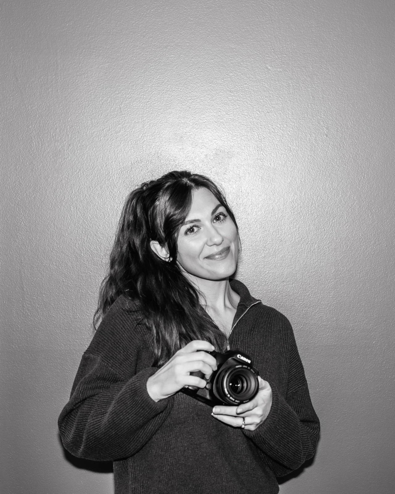 A woman with wavy hair holding a camera, standing against a plain wall, smiling at the camera.
