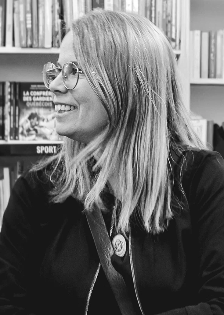 Une femme souriante portant des lunettes et une veste noire, assise dans une bibliothèque.