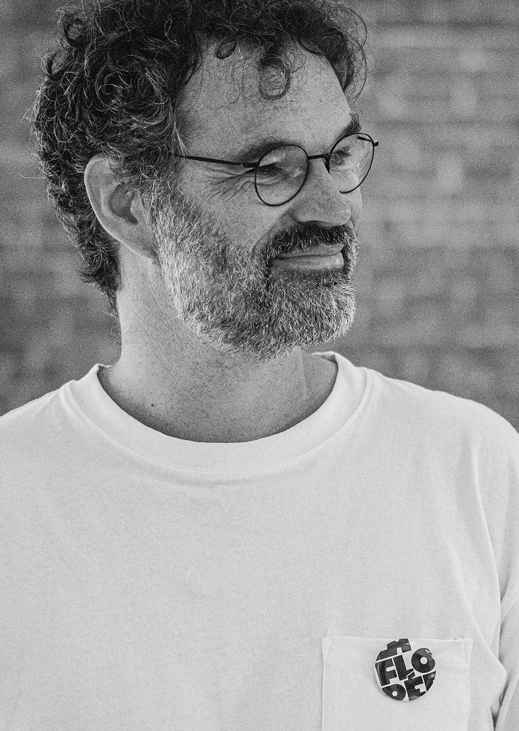 Un homme barbu portant des lunettes, vu de profil, souriant, portant un t-shirt blanc avec un bouton sur la poche. Fond flou de briques.