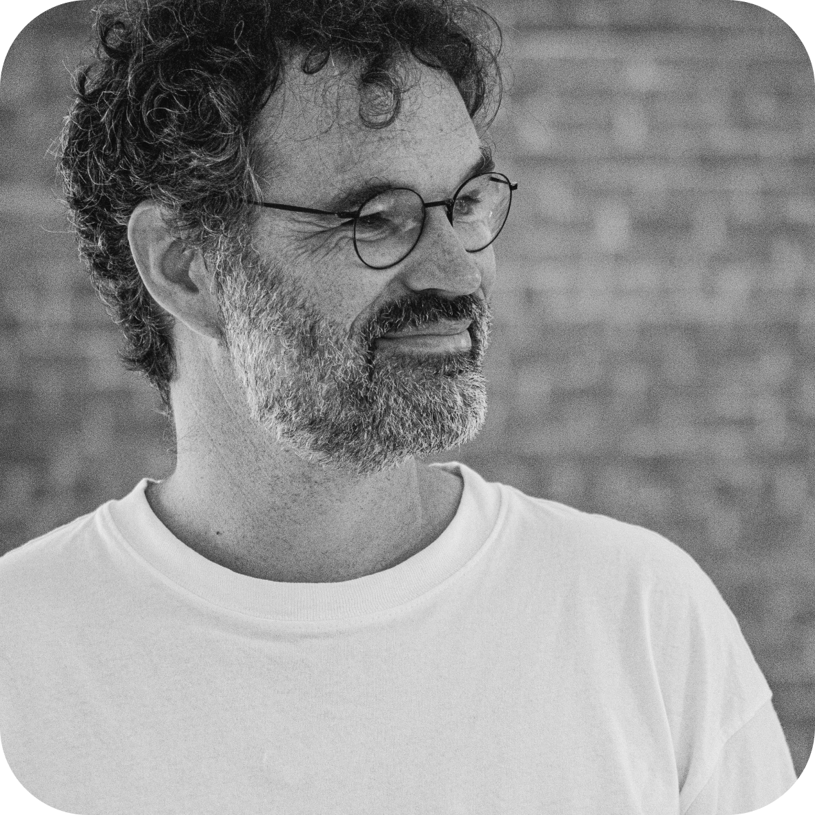 Homme avec des cheveux bouclés, une barbe grise, portant des lunettes rondes et un t-shirt blanc, souriant doucement.