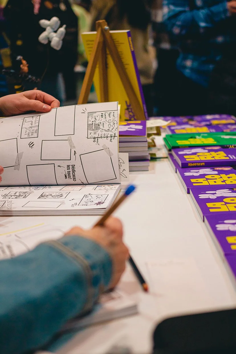 Une personne écrit dans un cahier à côté d'une table avec des livres violets et verts empilés, et un tableau de dessin sur un chevalet.