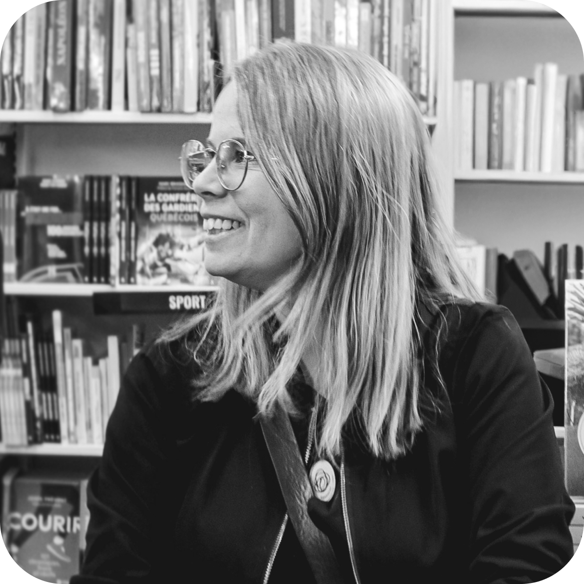 Une femme souriante portant des lunettes ronde assise dans une librairie, entourée de rayonnages remplis de livres.