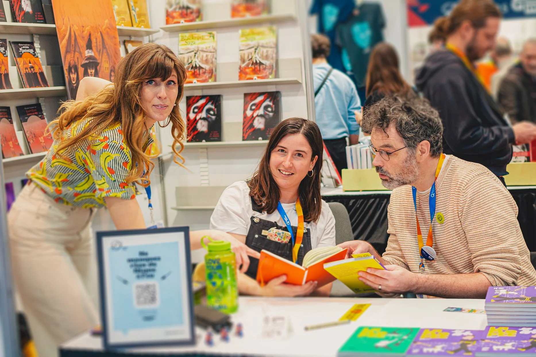 Trois personnes à une table de signature de livres dans une librairie lors d'une foire du livre, entourées d'étagères avec des livres et des visiteurs en arrière-plan.