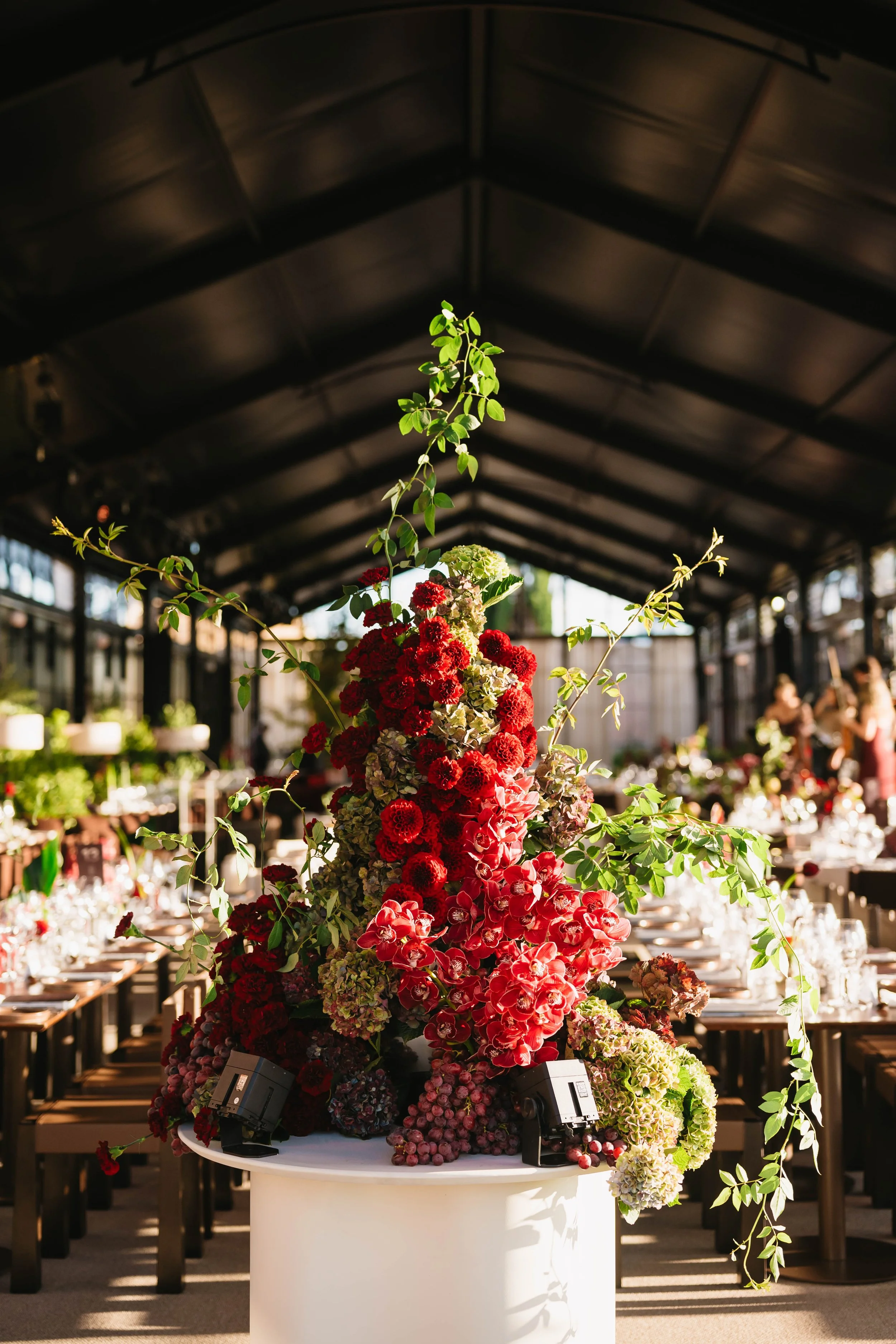 A floral arrangement with red, pink, and green flowers and foliage, set on a white table in a brightly lit indoor space with dark ceiling and large windows in the background.