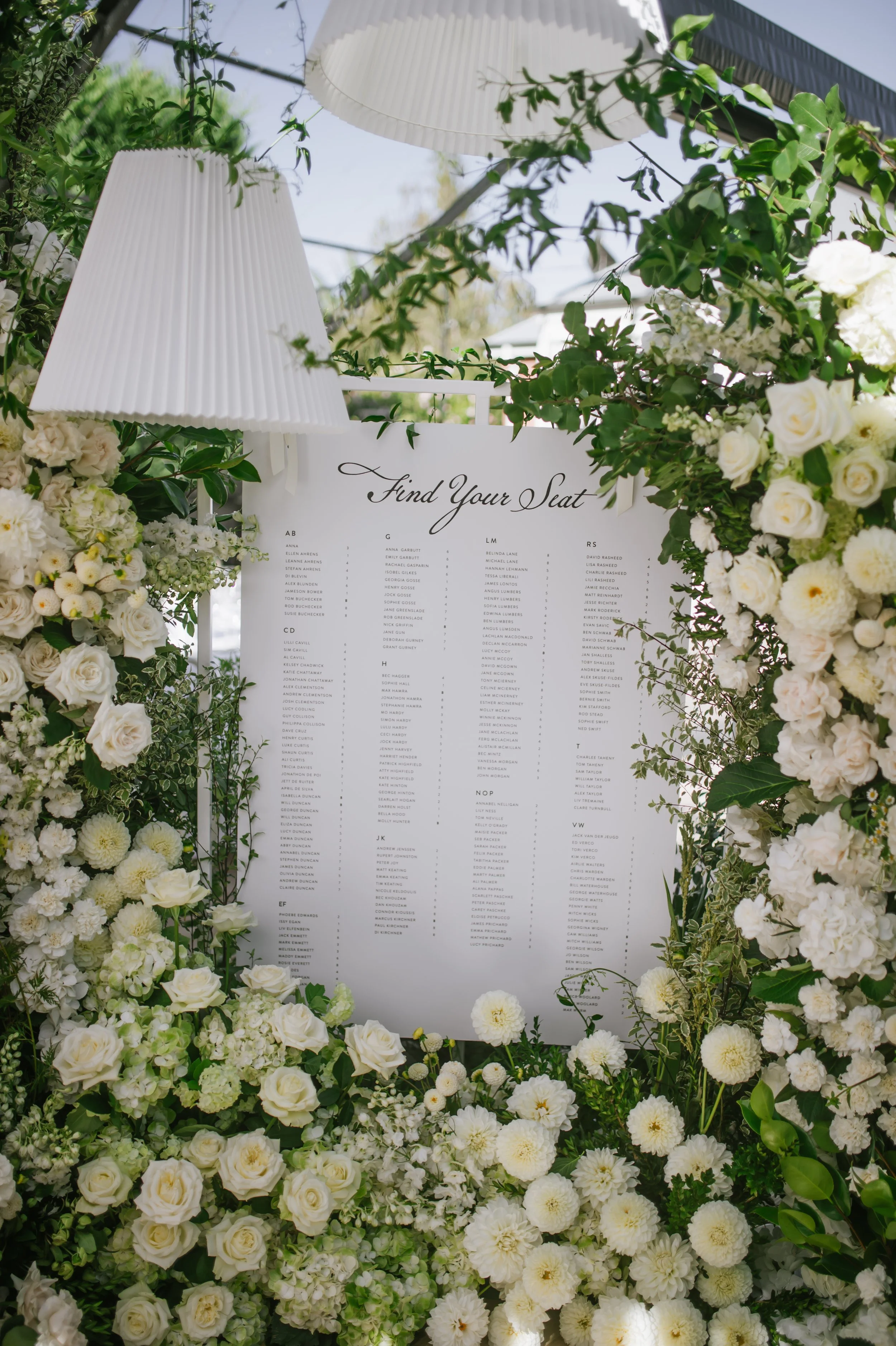 A floral wedding seating chart display surrounded by an abundance of white flowers and greenery, with a white lampshade hanging above.