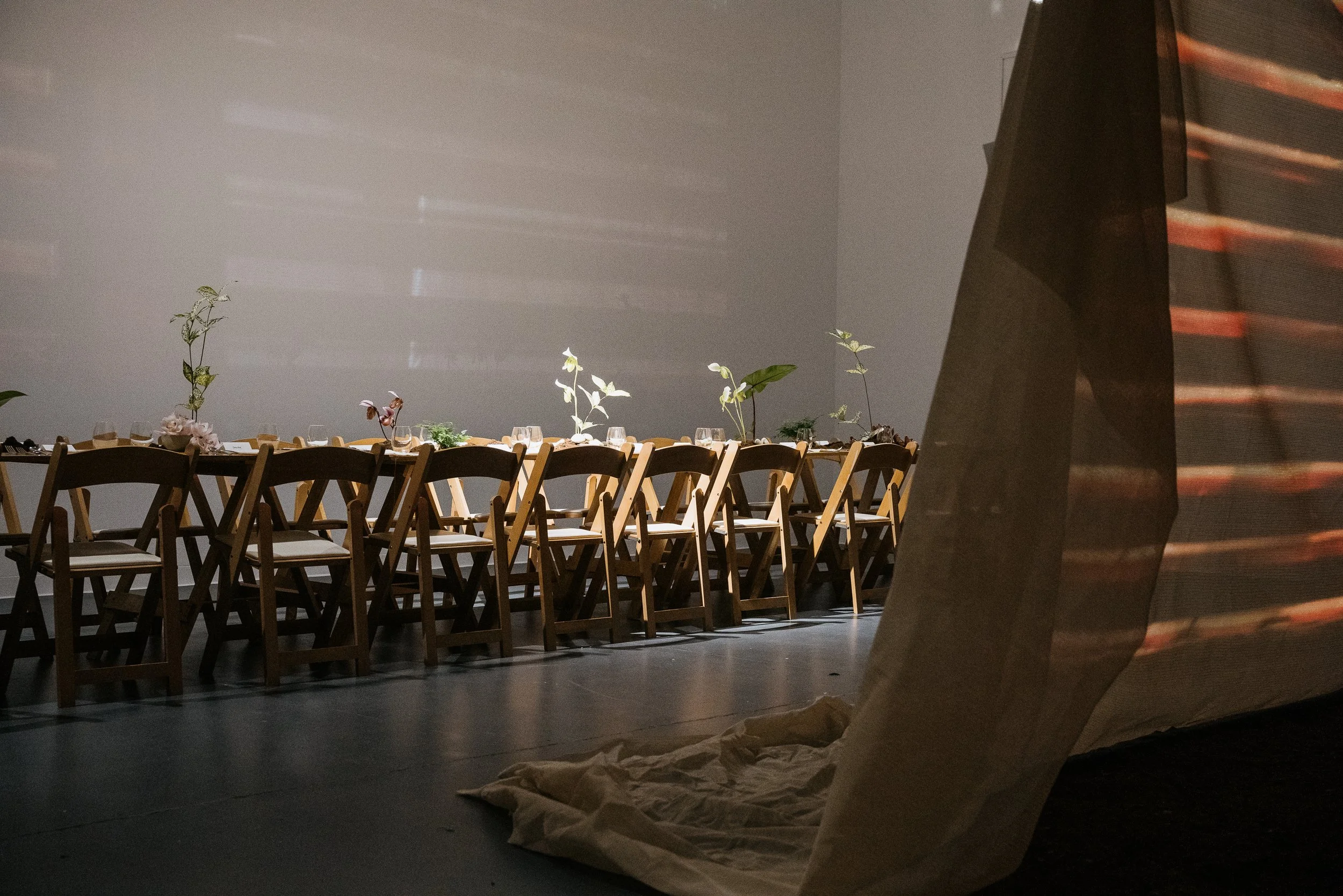 Interior view of a dimly lit banquet or event space with a long table set with glassware and decorated with tall floral arrangements. In the foreground, there is a large paper bag or gift bag on the floor, partially visible. To the right, a large bla
