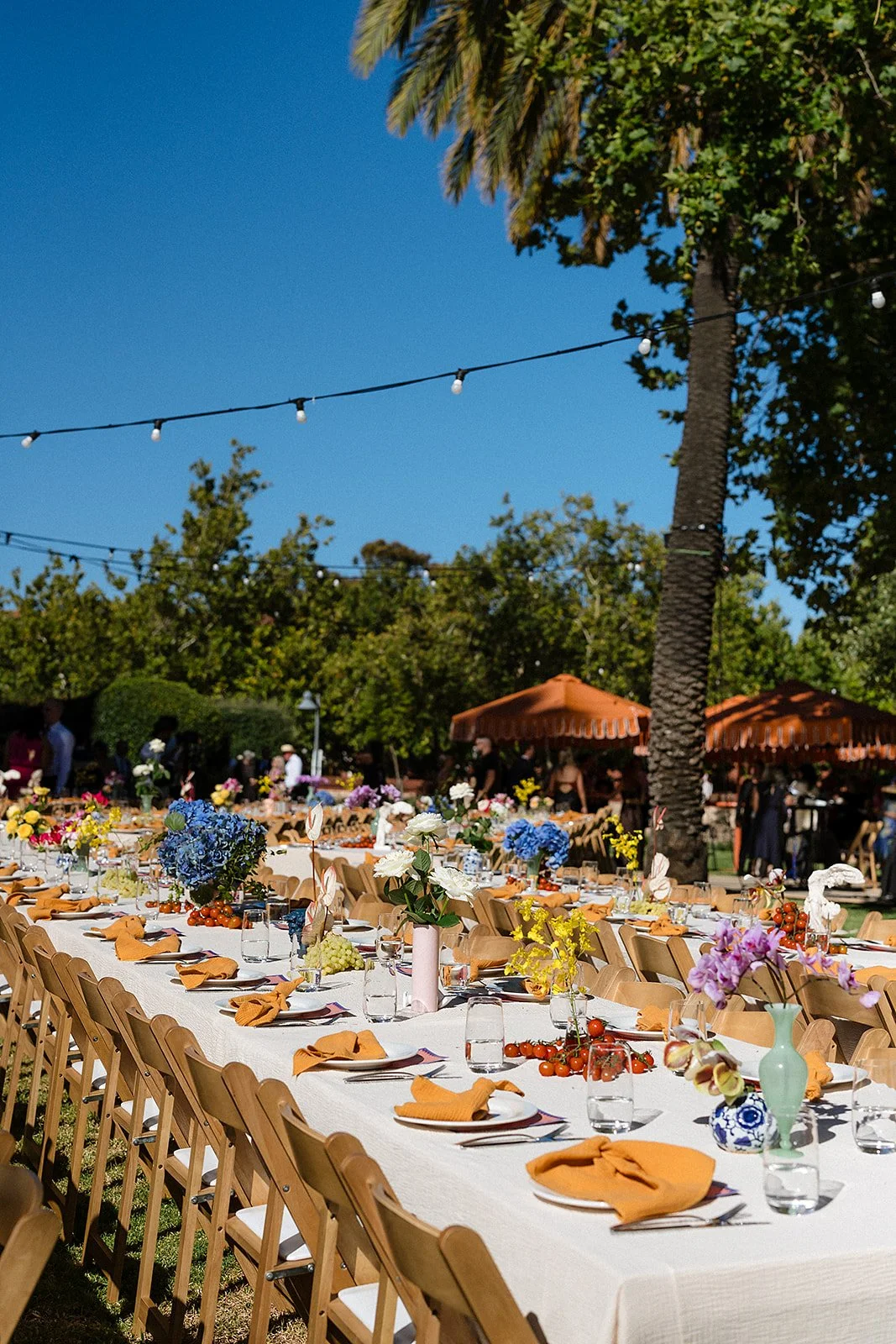 Long outdoor banquet table set with orange napkins, glass vases filled with colorful flowers, and small clusters of cherry tomatoes, under string lights and surrounded by trees and umbrellas.