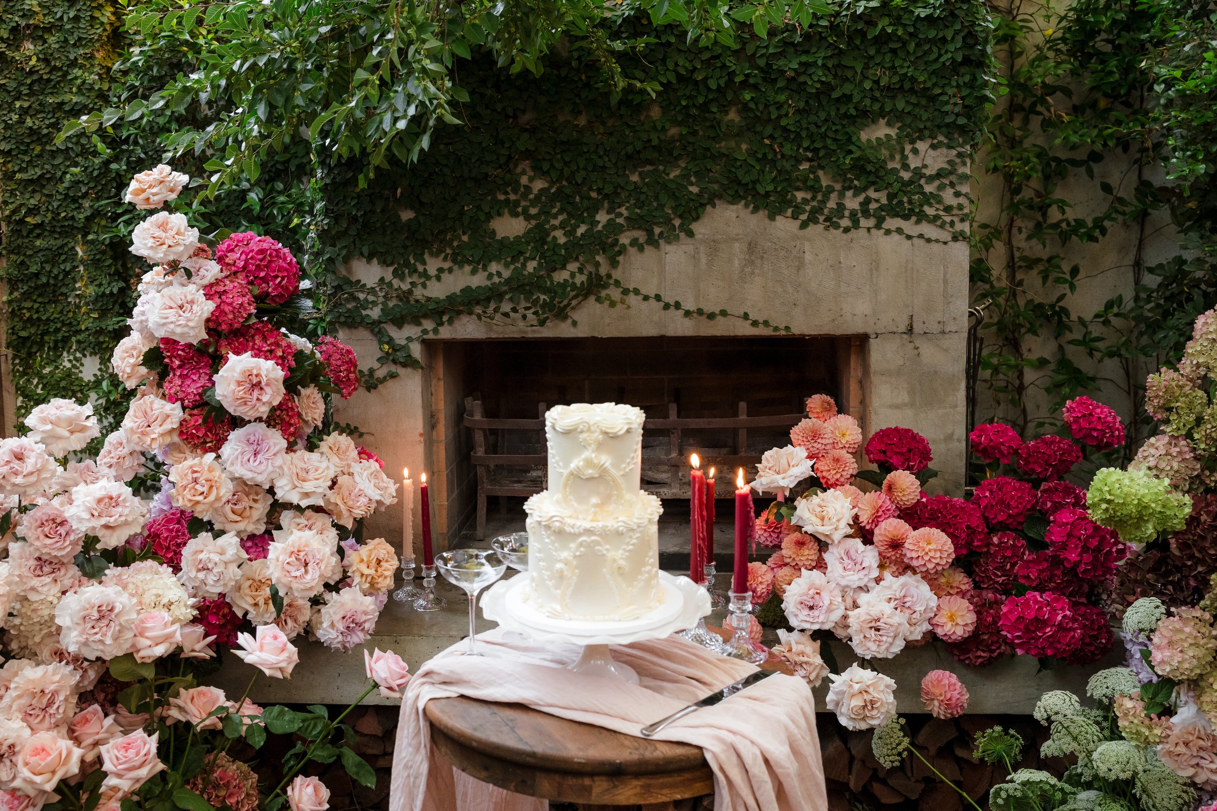 A decorated table with a three-tiered white wedding cake, surrounded by pink and white flowers, candles, and a pink cloth, set in front of a stone fireplace with greenery and ivy on the wall.