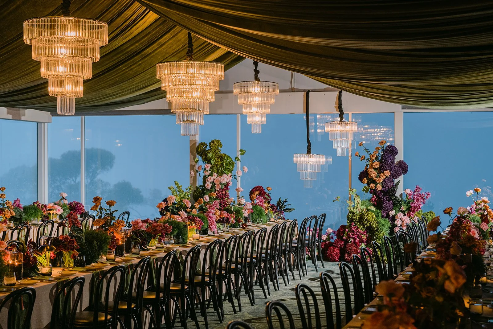 Elegant banquet hall decorated with multiple floral arrangements, chandeliers hanging from a draped ceiling, and large windows showing a cloudy sky outside.