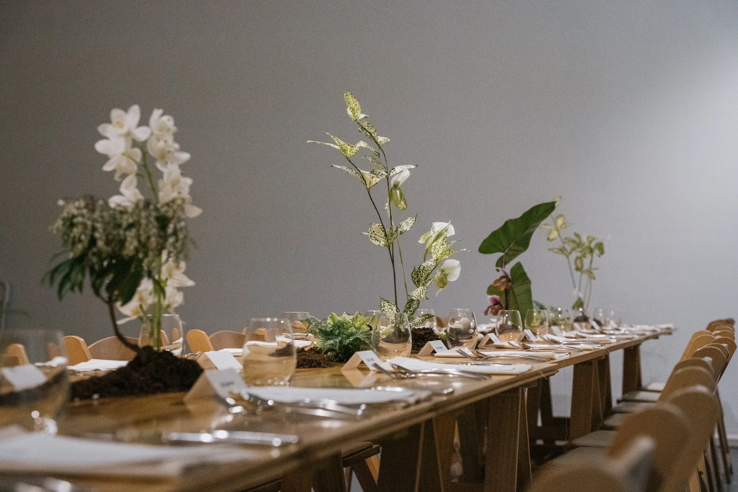 A long table arranged for a formal dinner with elegant floral centerpieces and place settings, including glasses and napkins, in a room with plain gray walls.