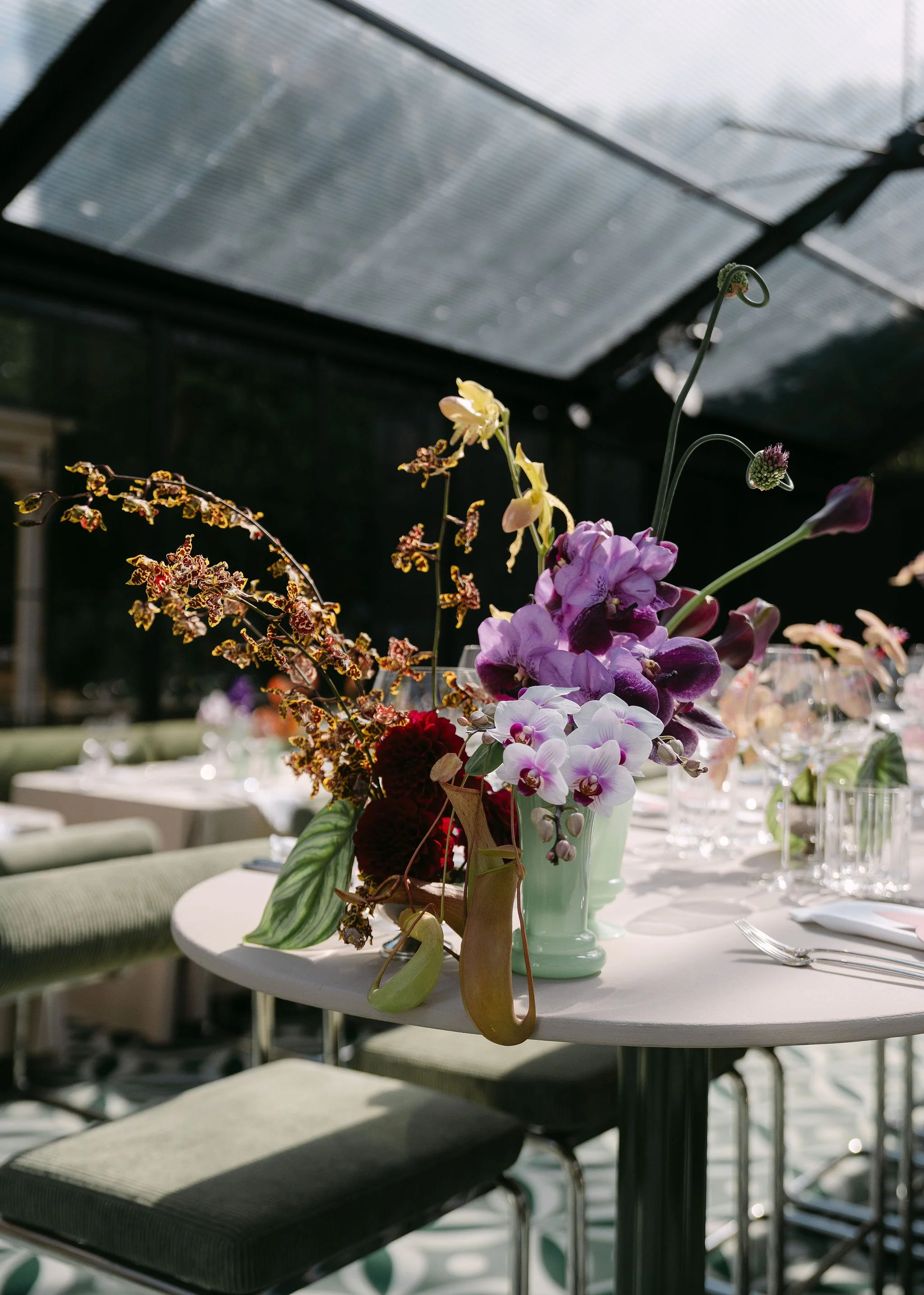 A close-up of a floral centerpiece on a white dining table, with a green vase holding purple, pink, and yellow flowers, set in a glass-enclosed outdoor space with chairs and tables in the background.