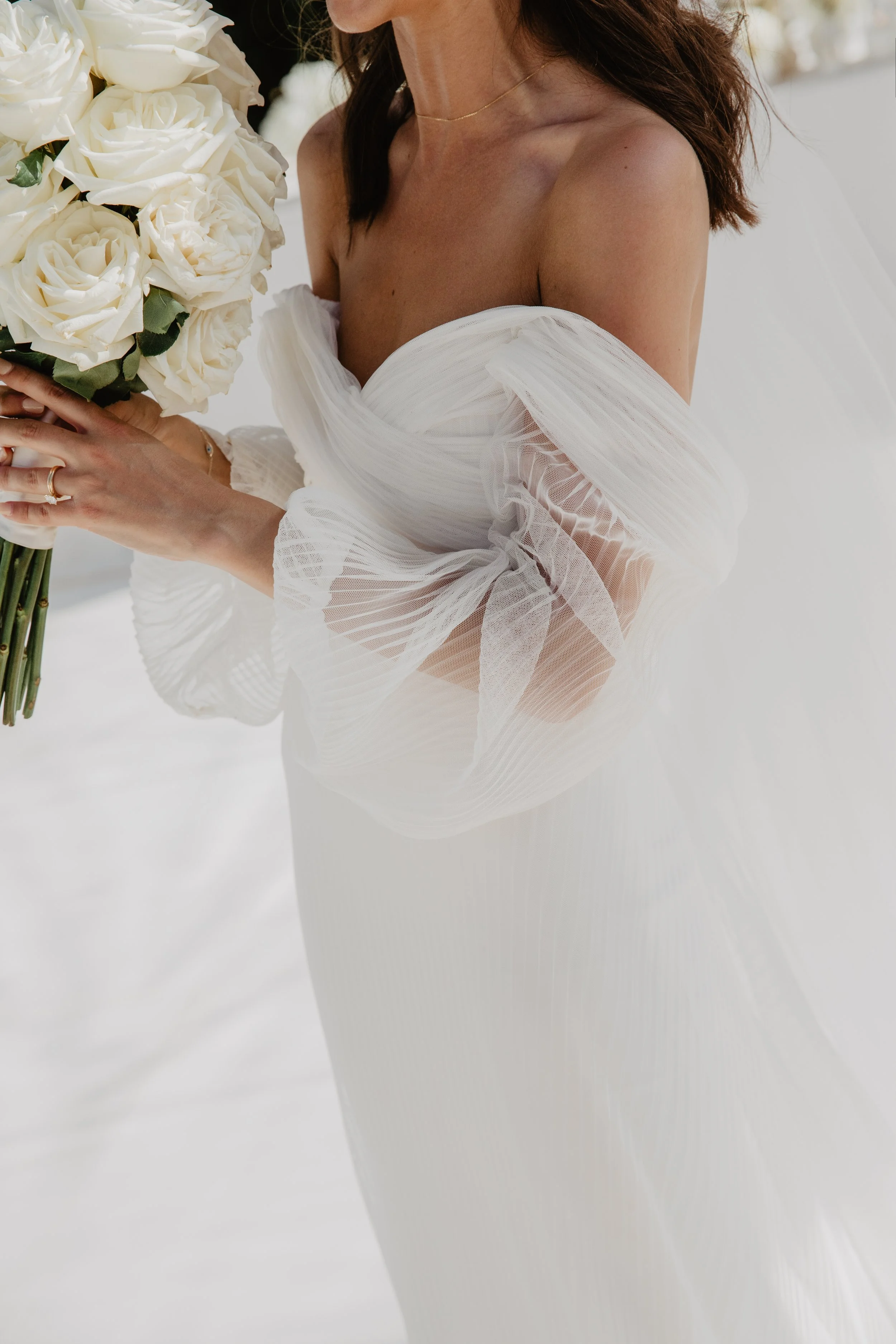 A bride in a white wedding dress holding a large bouquet of white roses. Part of her face is not visible, focusing on her shoulder, hand, and the bouquet.