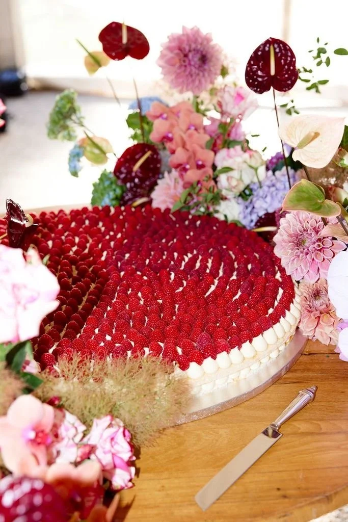 A cake decorated with raspberries, surrounded by pink and purple flowers and green foliage.