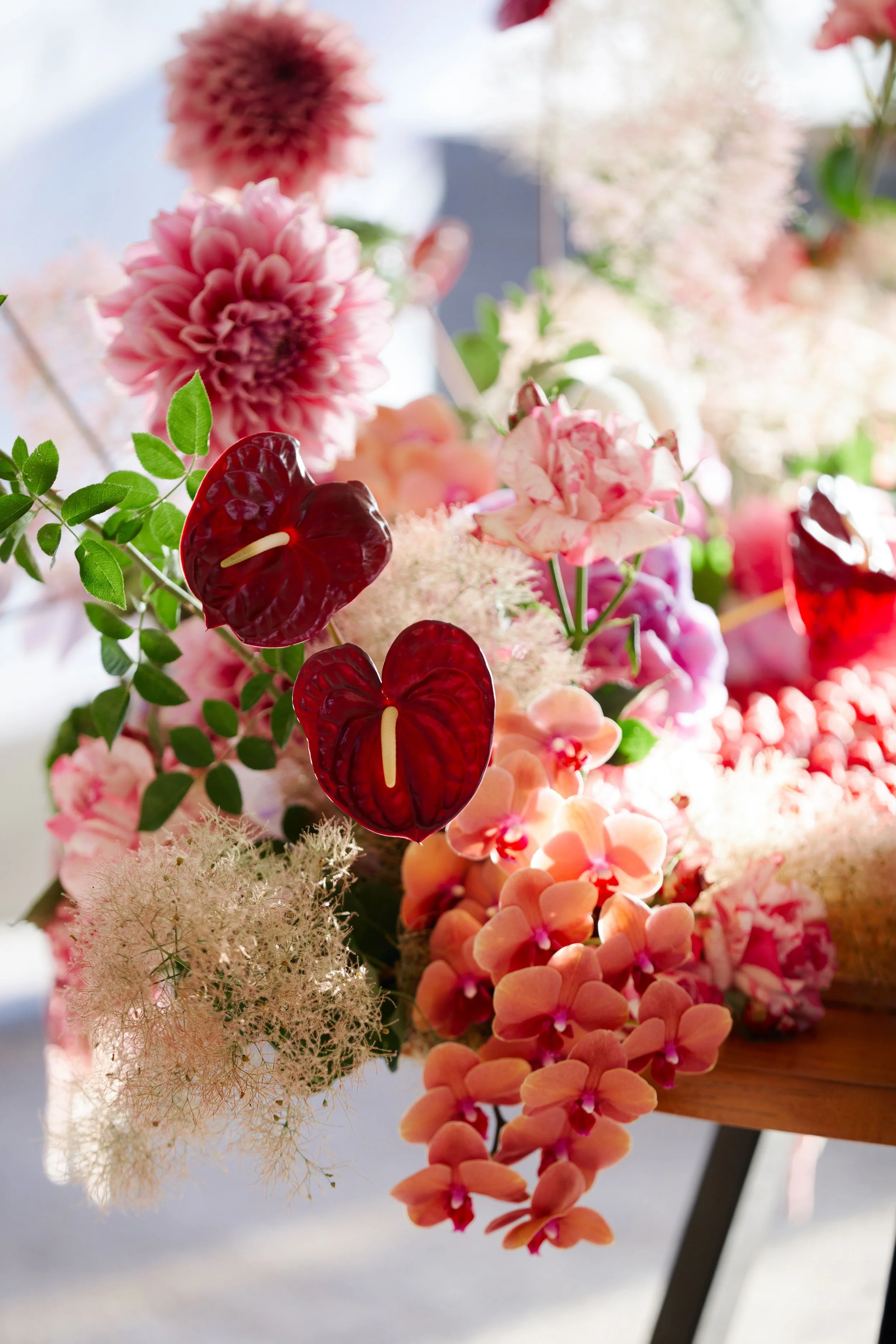 Close-up of a floral arrangement with pink dahlias, red anthuriums, peach and pink orchids, and white fluffy flowers, with green leaves and a wooden surface.