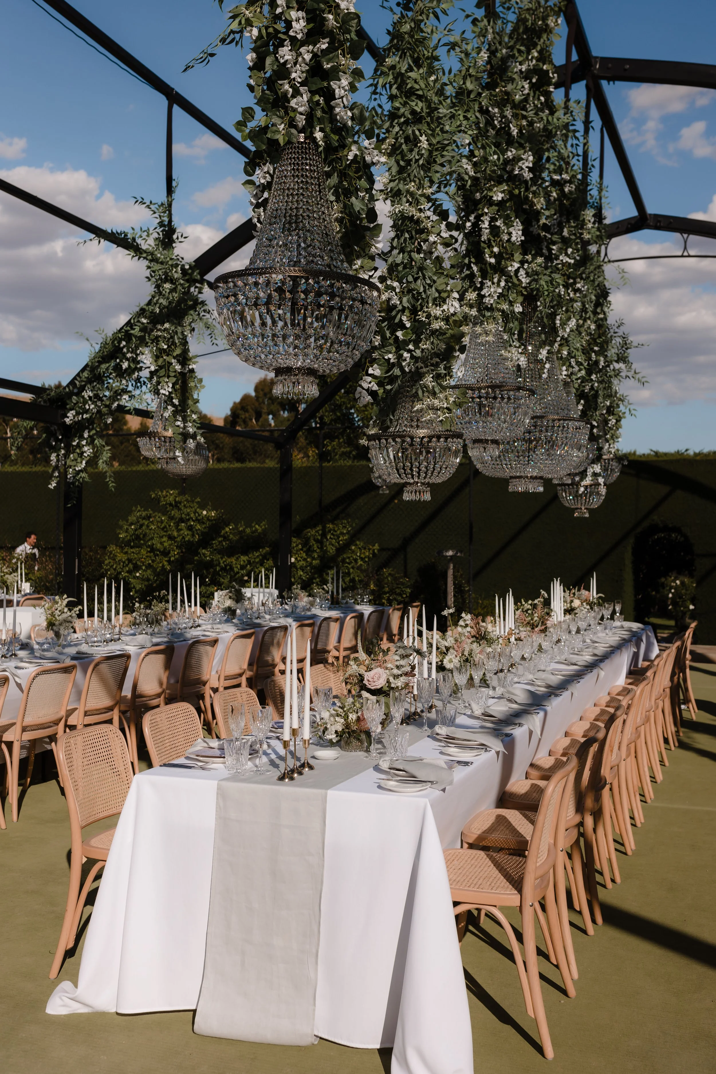 Elegant outdoor dining setup with long tables covered in white tablecloths, decorated with floral centerpieces, candle holders, and glassware, under crystal chandeliers and greenery decorations.