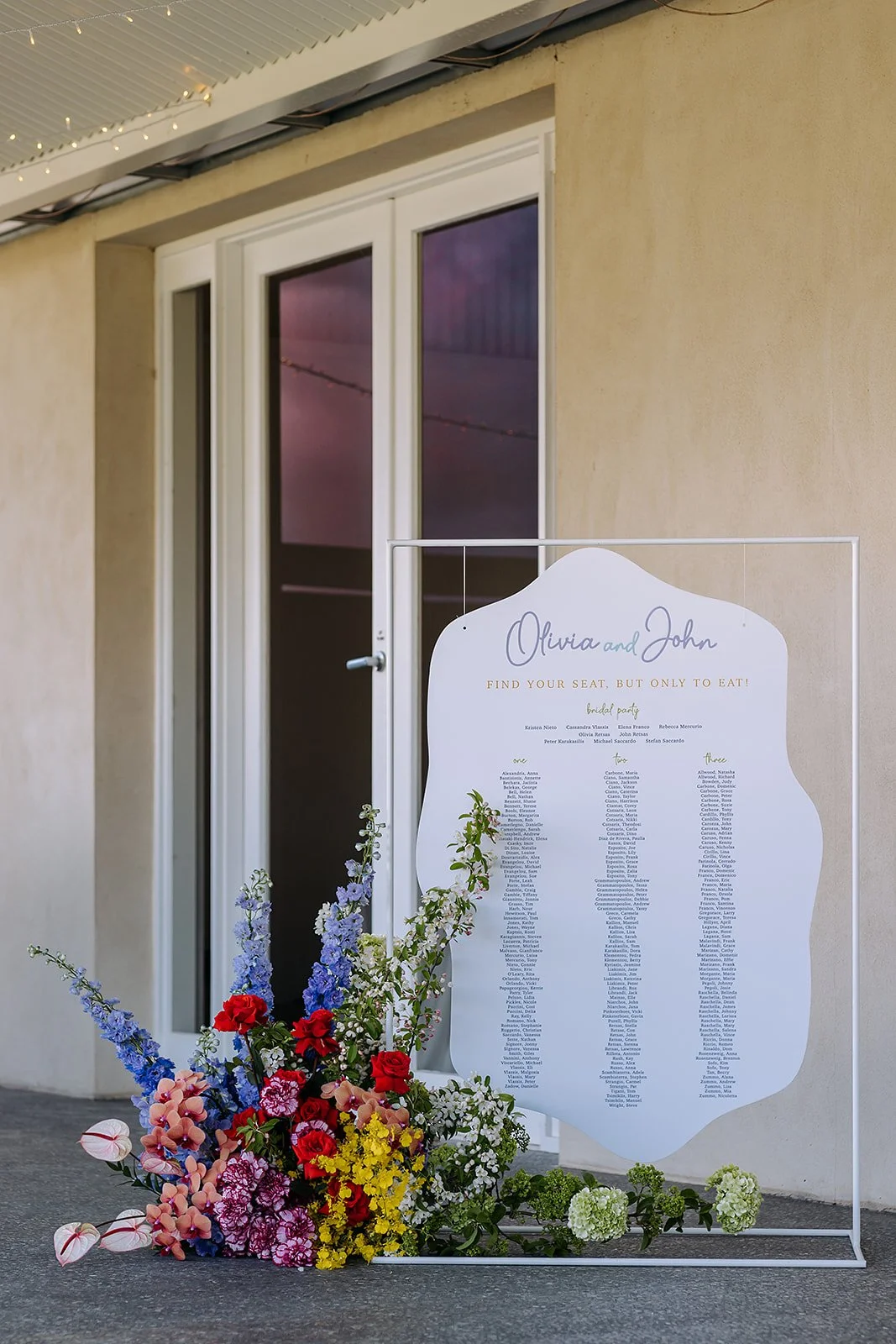 Wedding seating chart with colorful flower arrangement in front, placed outside near a beige wall and glass door, with string lights overhead.