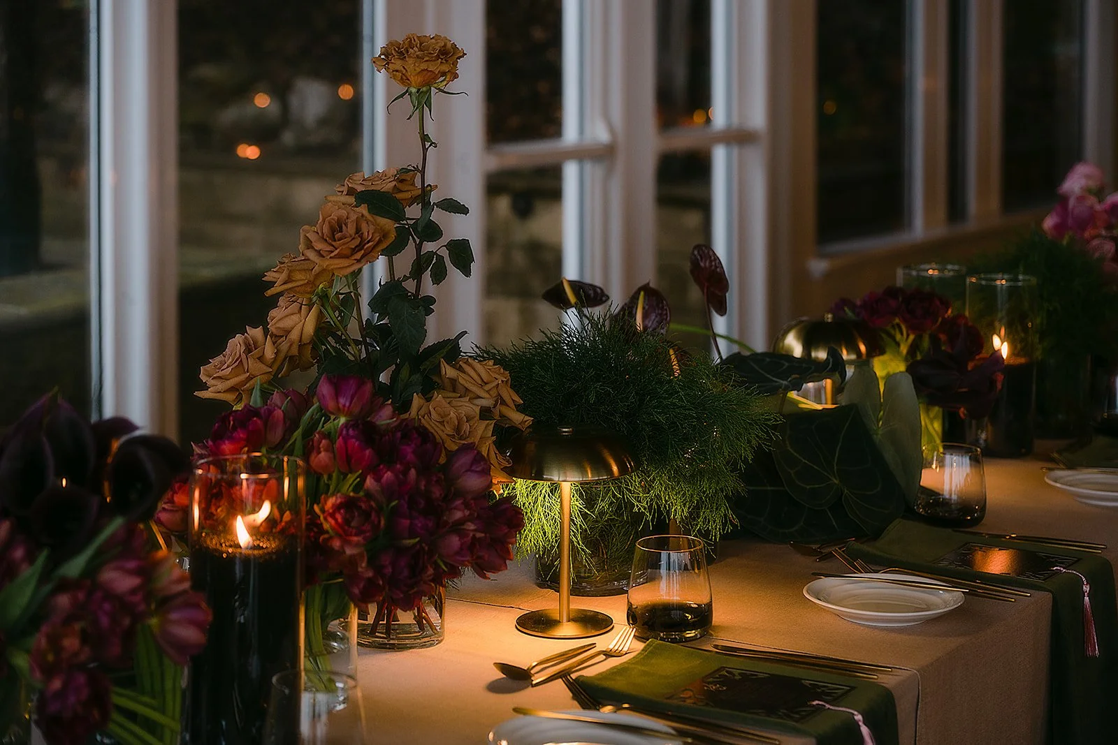 A decorated dinner table with flowers, candles, and tableware inside a room with large windows at night.