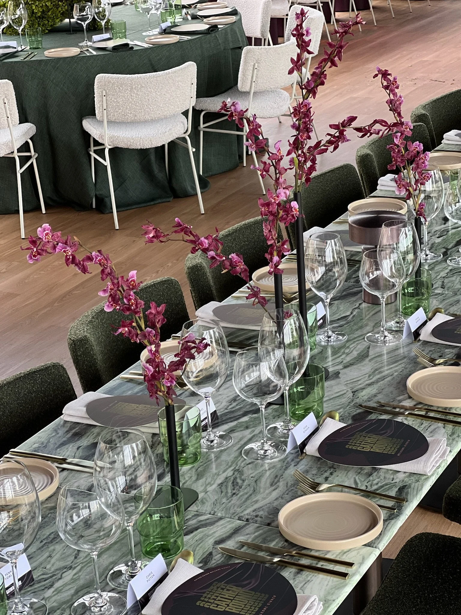 A long banquet table set for a formal event with pink flowers in tall black vases, wine glasses, green water glasses, beige plates, gold cutlery, and menu cards on a marble tabletop. In the background, there are white chairs and a table with a dark g