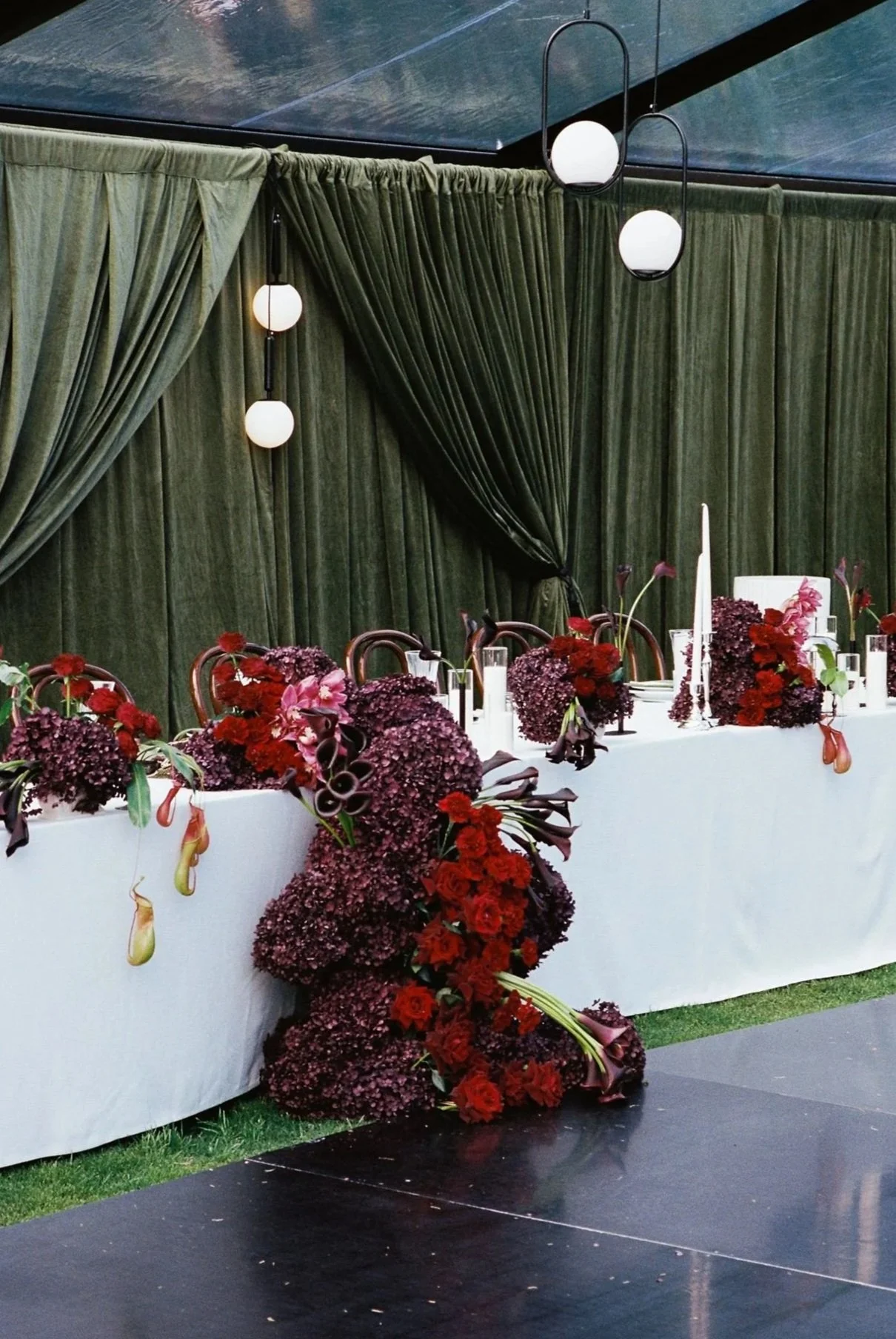 Elegant banquet table decorated with large pink and purple floral arrangements, white tablecloth, white candles, surrounded by wooden chairs, set against a dark green curtain backdrop with modern black and white pendant lights hanging from the ceiling.