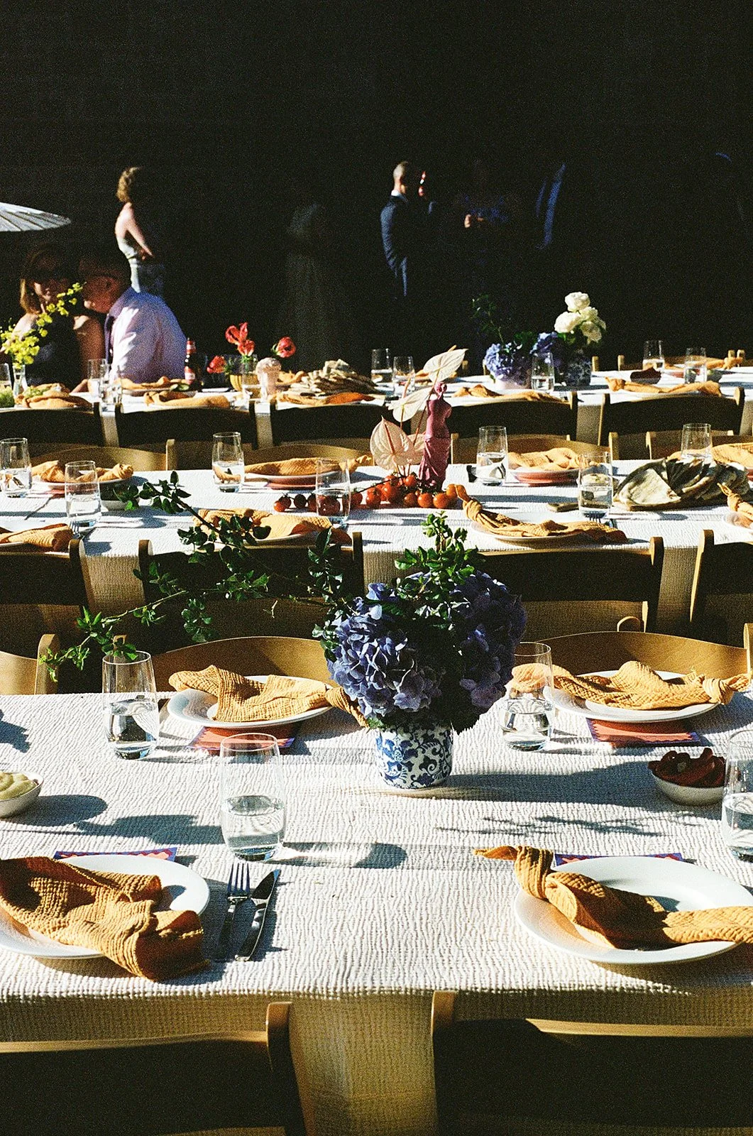 A decorated banquet table set with plates, glasses, napkins, and floral centerpieces at an outdoor event, with people socializing in the background under sunlight.