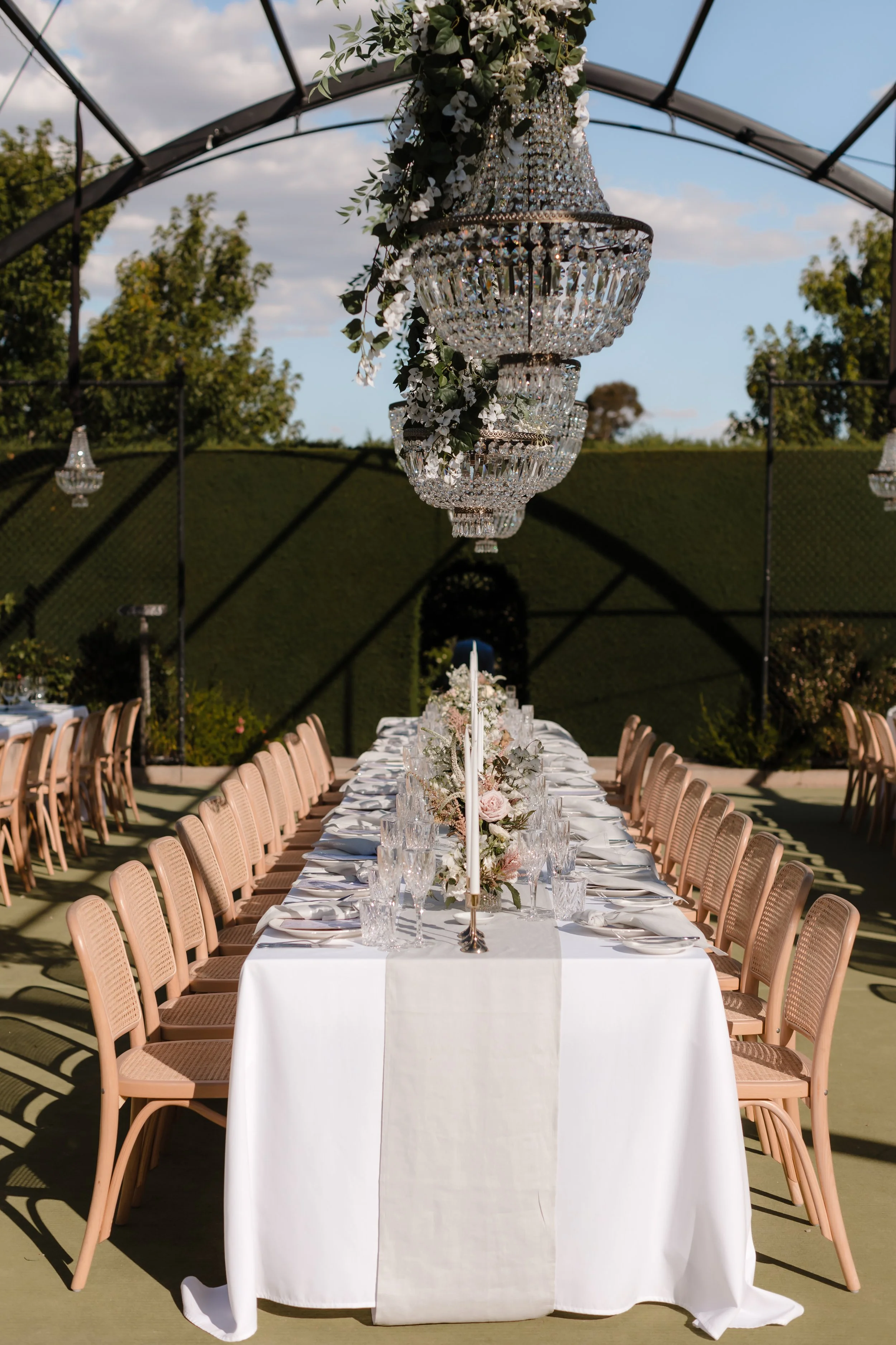 Elegant outdoor dining setup with a long rectangular table draped in white tablecloth, floral centerpiece, and tall white candles in silver holders, under a canopy of chandeliers, against a green hedge and blue sky.