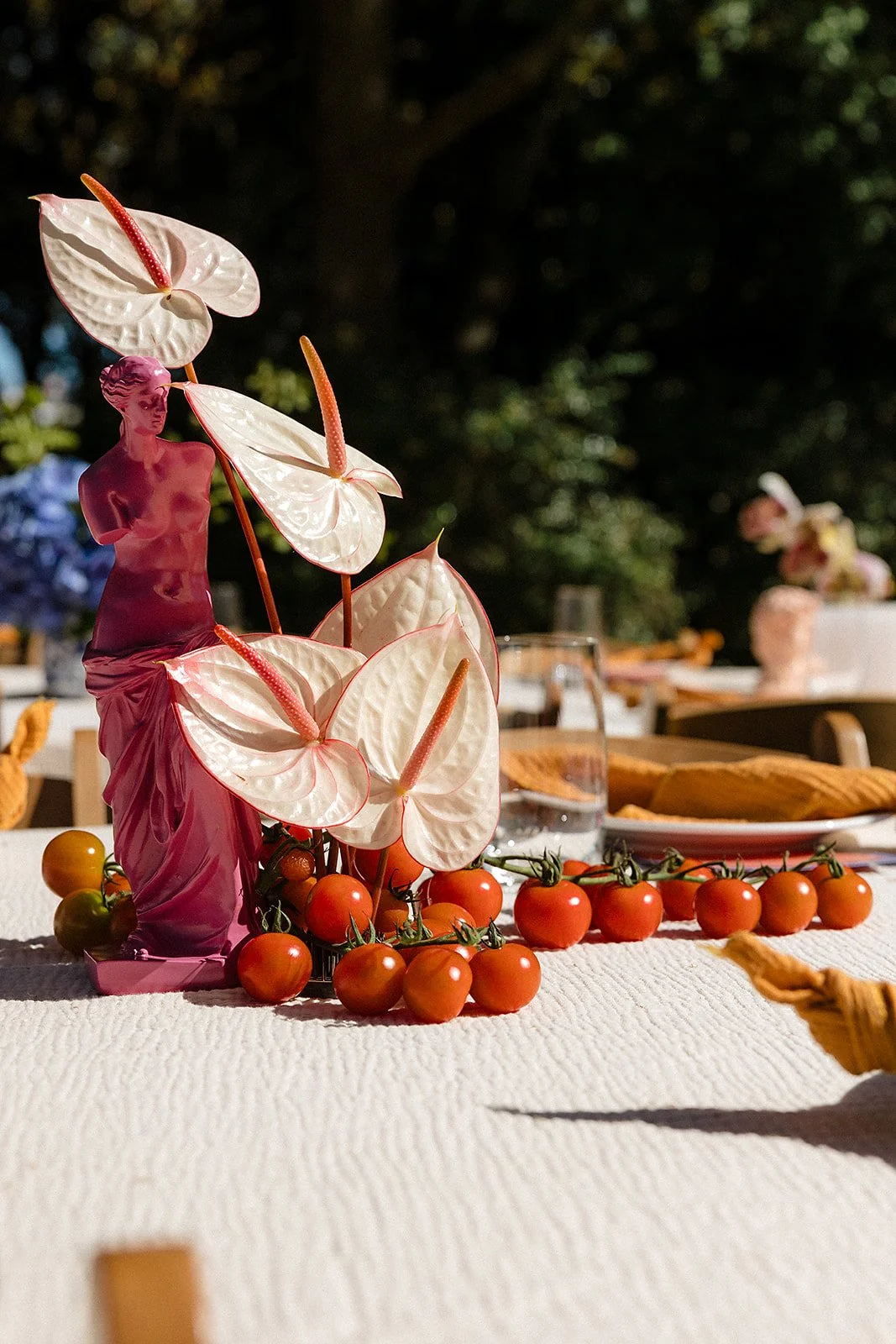 Decorative arrangement with cherry tomatoes, an artificial pink female statue, and large white artificial anthurium flowers on a table, outdoors in a garden setting.