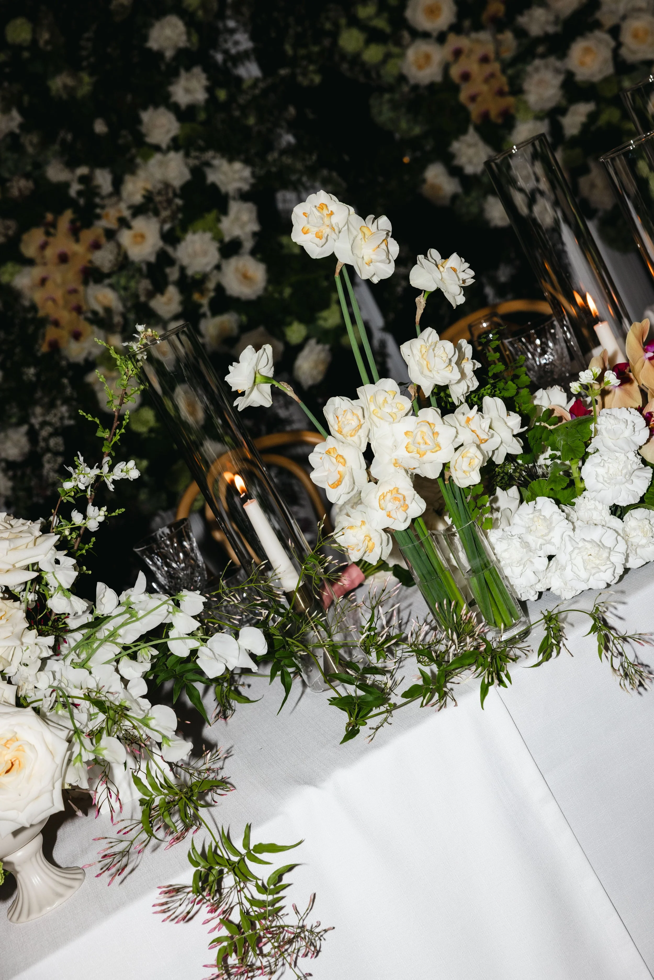 Elegant floral centerpiece with white and cream flowers, surrounded by glass candle holders with lit candles, on a white tablecloth.
