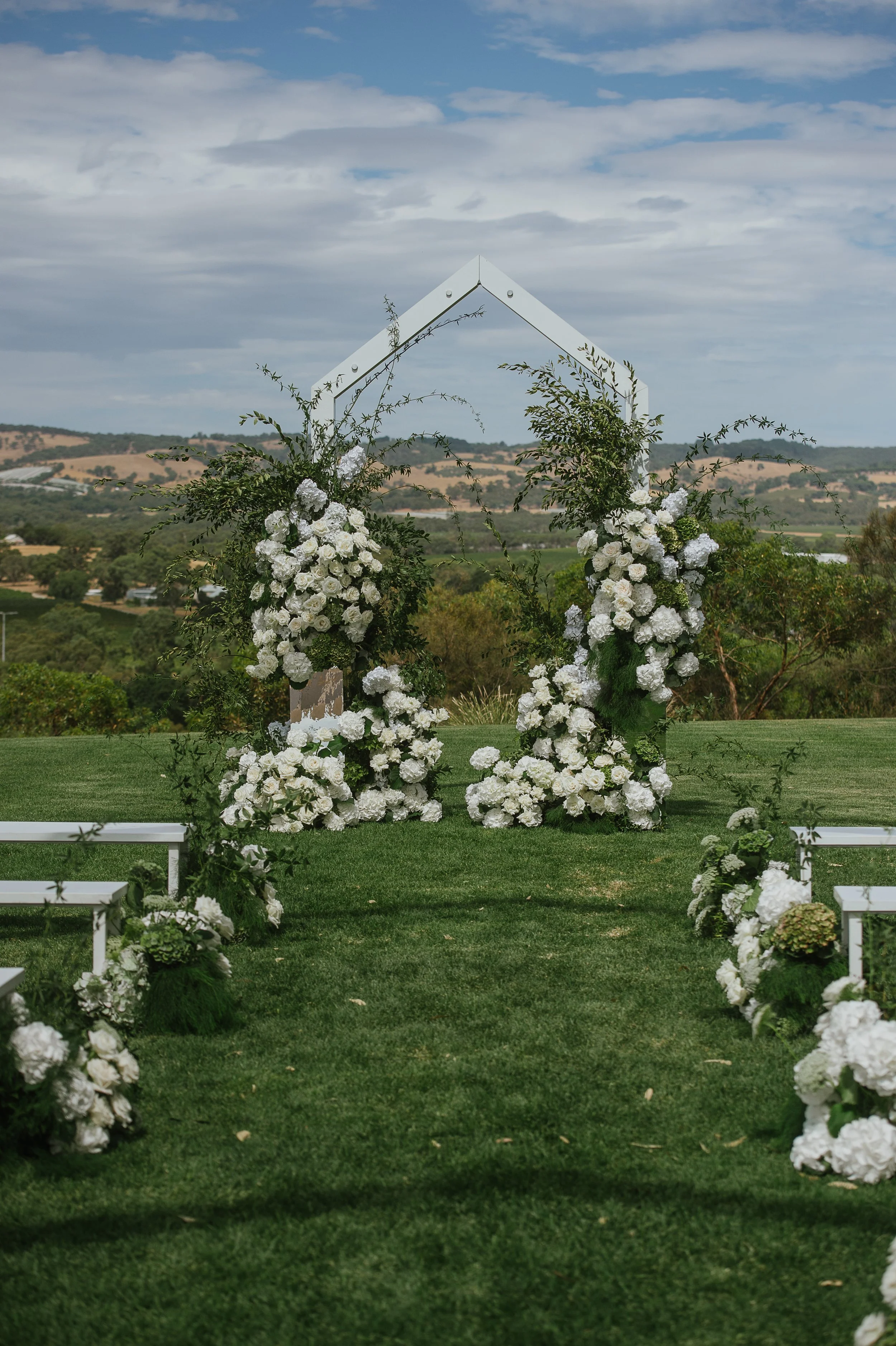 Outdoor wedding arch decorated with white flowers and greenery, set on a grassy area with a scenic rural landscape background under a partly cloudy sky.